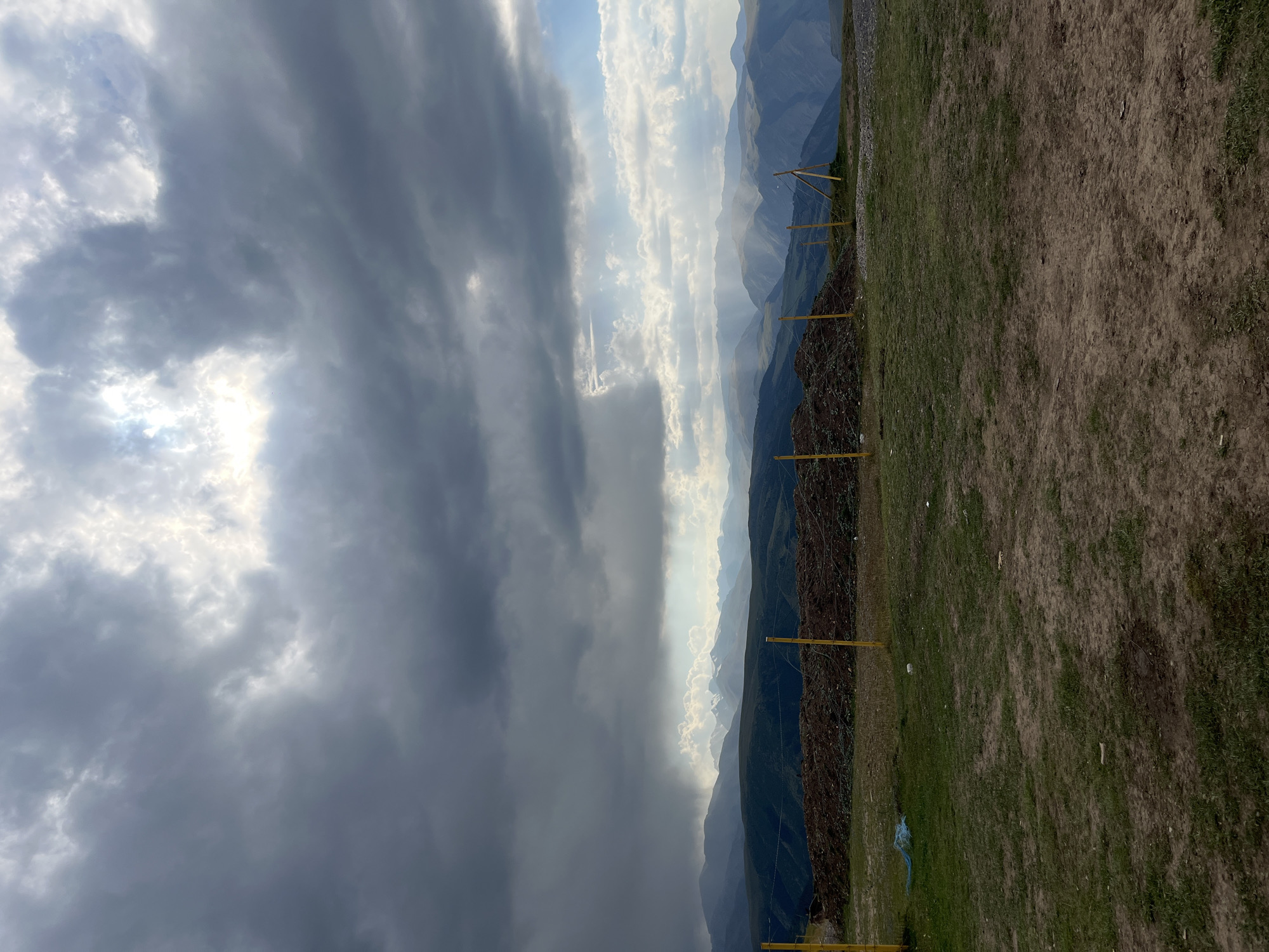 The image shows a panoramic view of a mountainous landscape under a dramatic, cloudy sky. The foreground consists of dry, patchy grass with some dirt. A mound of dark, rocky material is piled behind a fence made of yellow poles and barbed wire. Beyond this are several layers of rolling hills and mountains, receding into the distance. The sky is filled with heavy, dark grey clouds, with breaks of sunlight creating dramatic shafts of light that illuminate parts of the distant mountains. A patch of light rain or mist is visible falling on the farthest mountain range. There are no people or animals visible in the image. The scene suggests a remote, natural environment, possibly experiencing unsettled weather. The location in the city of Batan, Kazakhstan is not visually discernible from the image itself.