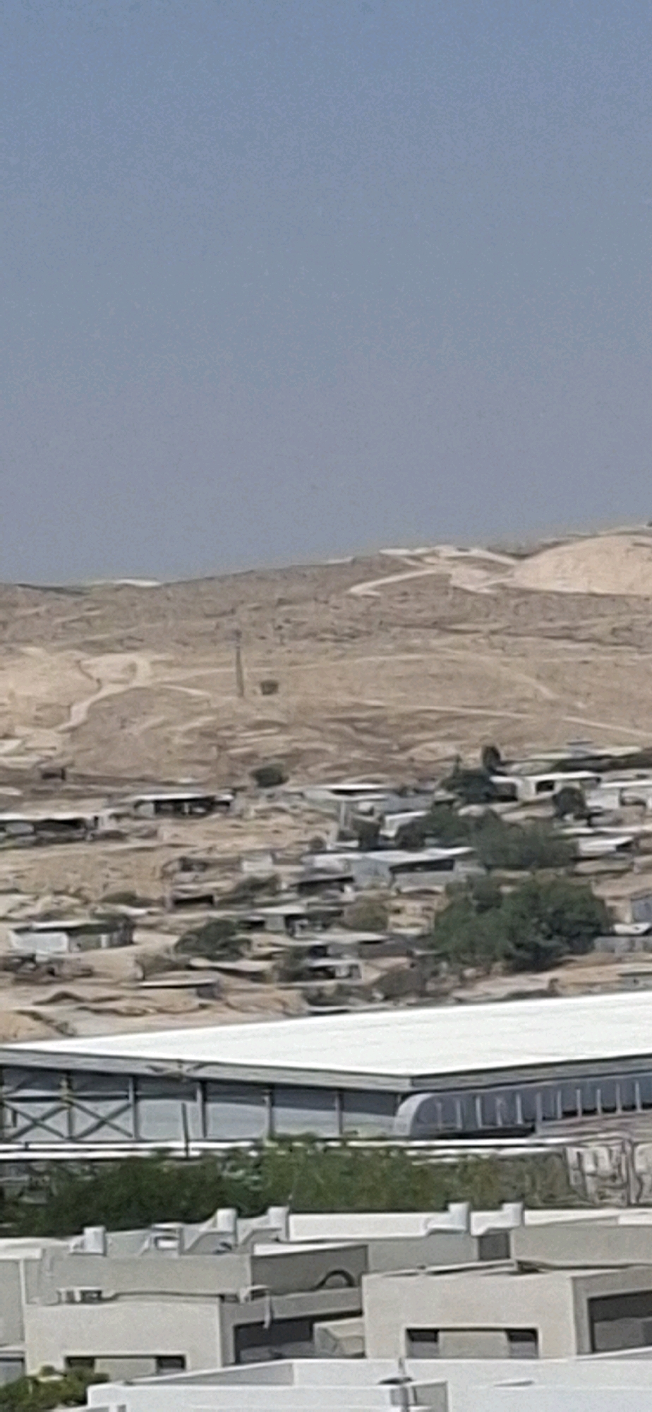 This is a photograph of a semi-arid landscape near Yerocham, Israel. In the foreground, there are several modern, multi-story buildings with white and grey facades. Behind these buildings, there is a large structure with a white roof, possibly a stadium or industrial building, with exposed metal beams and a row of windows. Further in the background, nestled in a valley, is a cluster of smaller, single-story buildings with grey or white roofs, suggesting a residential or industrial area. The landscape rises into hills in the distance, characterized by sparse vegetation and eroded terrain. A tall, slender tower, likely a utility pole or communication tower, stands on one of the hills, with a dark rectangular object near its base. The sky is a pale, hazy blue. The overall impression is of a developed area within a desert environment.