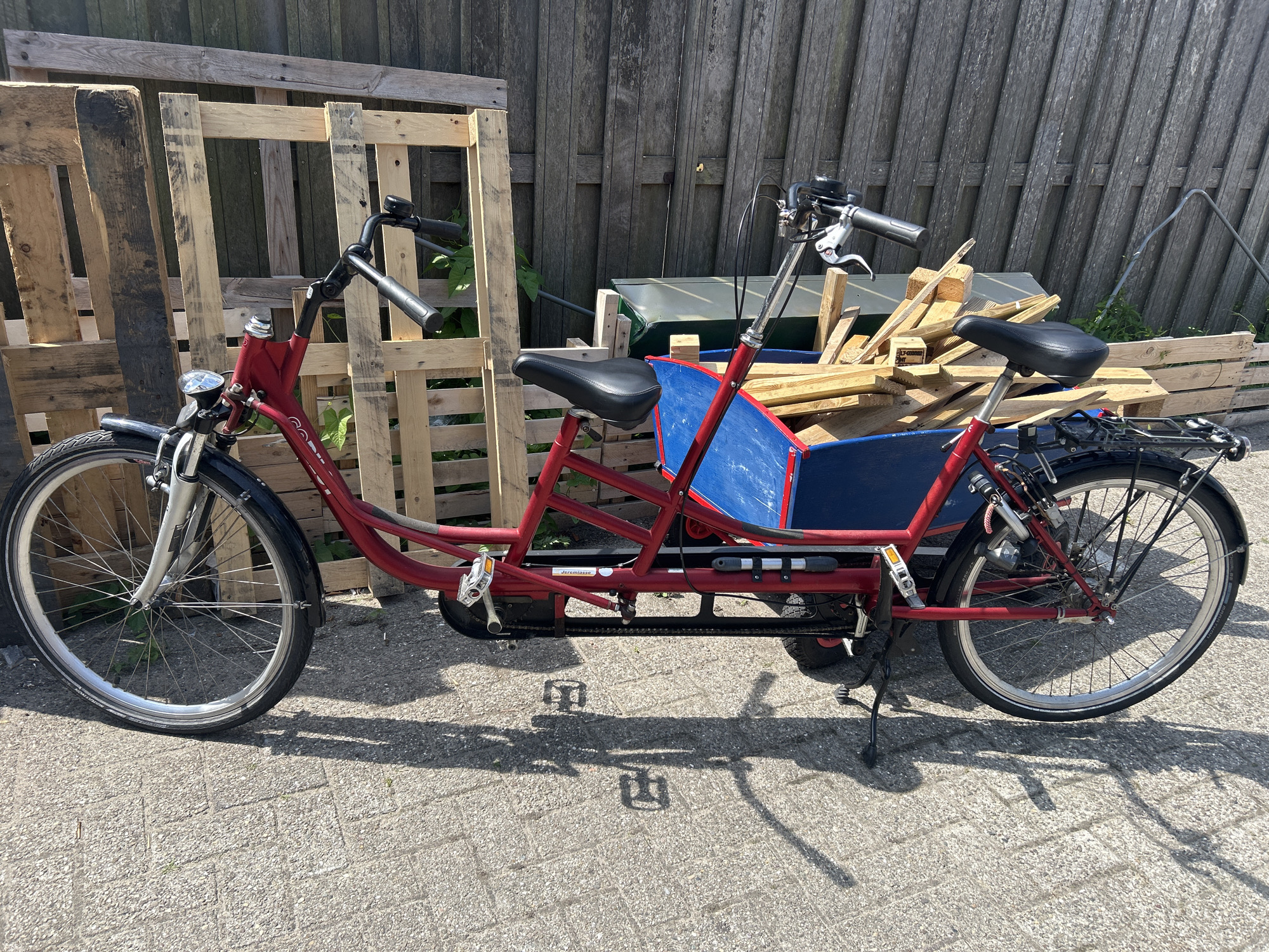 A red tandem cargo bicycle is parked outdoors on a paved surface, casting a shadow. The bicycle has two seats, handlebars for both riders, and a blue cargo box located between the seats. Behind the bicycle, a wooden fence and stacked wooden pallets are visible. The environment appears to be in Lisse, Netherlands.