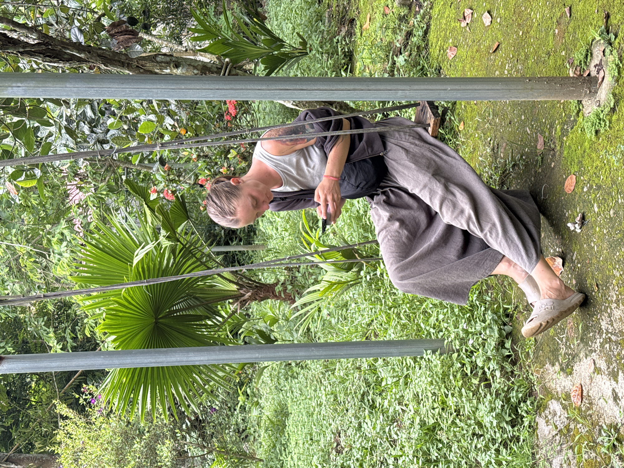 In Cẩm Thạch Commune, Vietnam, a woman with blonde hair tied back is seated on a wooden swing. She is wearing a grey tank top, a dark grey long-sleeved shirt, and wide-legged grey pants. A black fanny pack is worn across her chest. She is looking down at her phone, which she is holding in her hands. The swing is suspended between two metal poles, one of which is visible in the foreground. The background is lush and green, with various tropical plants, including palm trees and flowering bushes. The ground around the swing is covered in moss and scattered leaves.