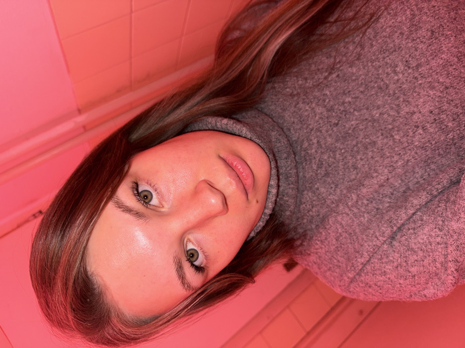 A young Caucasian female with long brown hair and green eyes is depicted in a close-up portrait, looking directly at the camera. She is wearing a grey, knitted turtleneck sweater. The scene is illuminated by a strong, uniform red light source, casting a crimson hue over the subject's skin, hair, and clothing, as well as the background. The background appears to be an interior wall, possibly tiled, showing faint horizontal and vertical lines indicative of grout or seams. The precise location within the city of Belgrade, Serbia, cannot be determined from the visual information provided in the image.