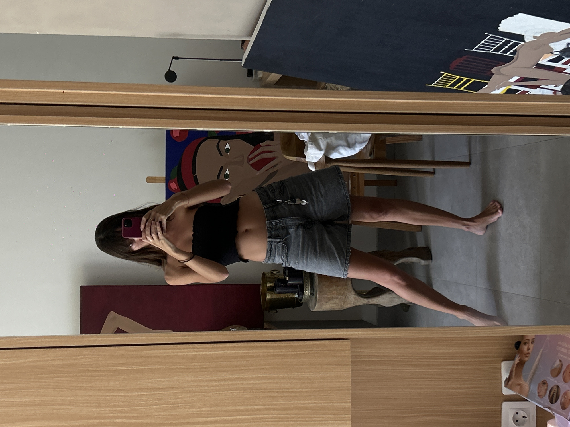 A woman is taking a photo of herself in a mirror. She is wearing a black top and denim shorts. She is standing in what appears to be a room in North Kuta, Indonesia, indicated by the electrical outlet. Behind her is a painting of a woman eating an apple, another painting with figures, a white towel, and various other objects on shelves.