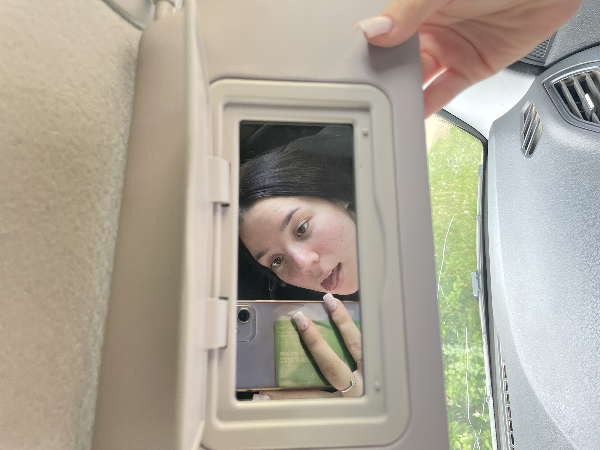 A woman is seen in a car, looking into a vanity mirror attached to the sun visor. She is holding a smartphone in her left hand, with a green card placed behind it. The woman is sticking her tongue out playfully. The mirror reflects her face, a portion of her hair, the smartphone, and the green card. Outside the car, green foliage is visible through the windshield. The sun visor is open, and a hand with painted nails is visible on the right side of the frame, adjusting the visor. The scene appears to be set in a natural outdoor environment, possibly a rural area or a park, given the abundant greenery. The location of Nozhovka, Russia is not directly ascertainable from the visual information provided.