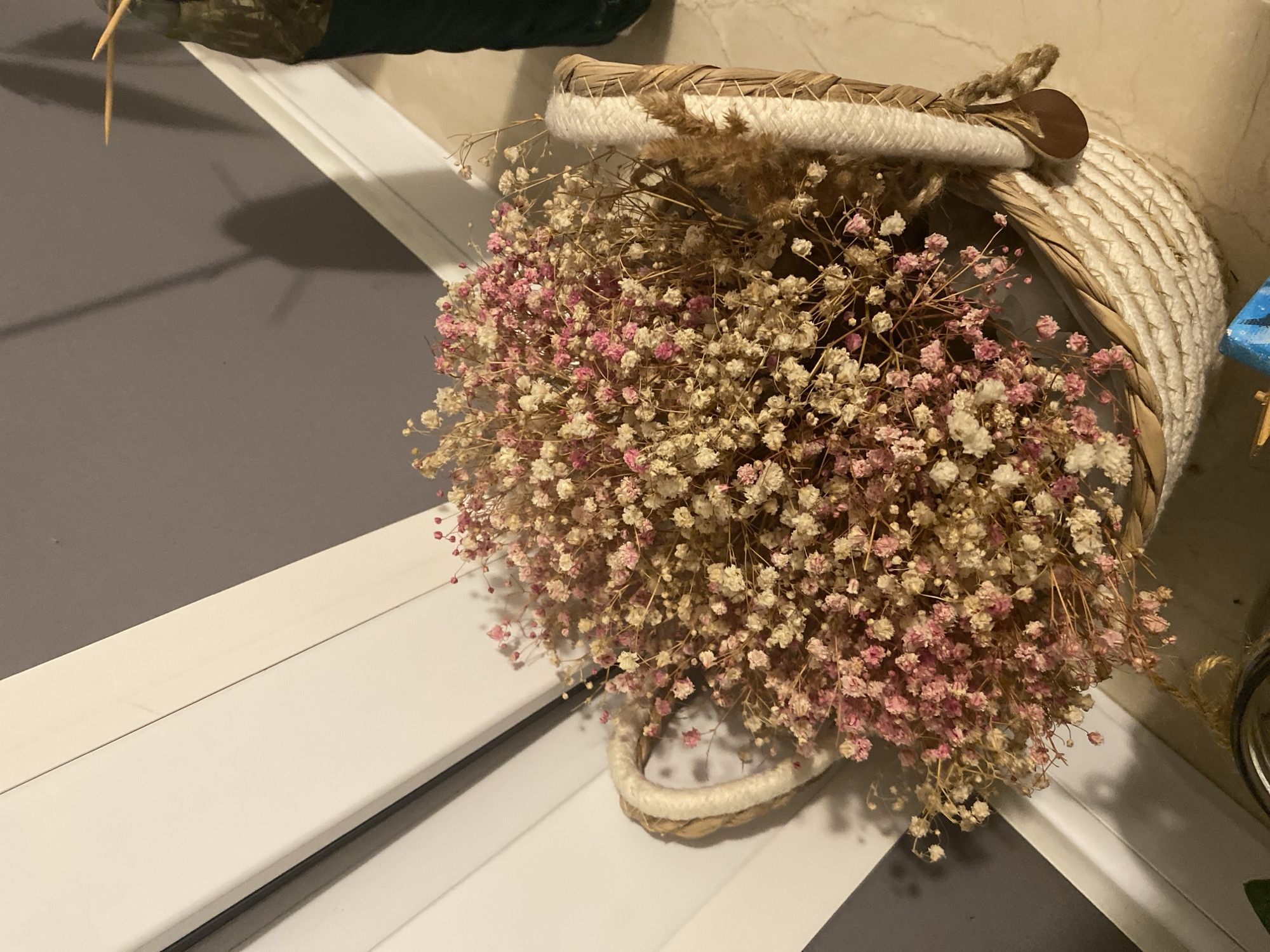 A woven basket filled with dried baby's breath flowers, some tinged with pink, sits on a marble floor next to a white trim that transitions into a grey wall. A shadow of a person is cast on the wall. The scene is indoors, likely in a residential or commercial space in Łódź, Poland. There are no people visible in the image.