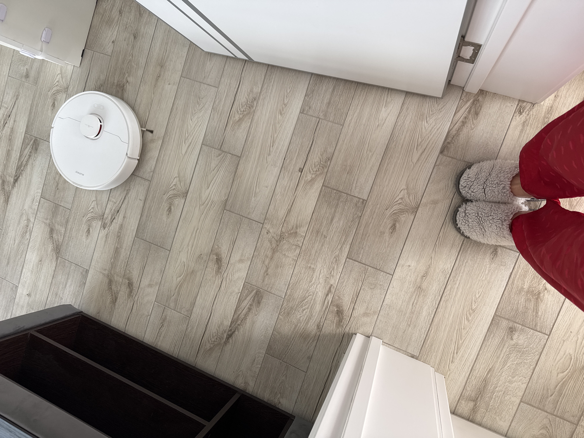 The image shows an indoor scene with light-colored wood-patterned flooring. A white robotic vacuum cleaner is positioned on the left side of the frame. Adjacent to the vacuum, a dark brown shoe cabinet or shelving unit is visible in the lower left corner. In the upper center and upper right, parts of a white door and adjacent wall are present. In the lower right corner, a pair of grey fluffy slippers and red pajama pants are visible on a person's feet. The specific location in the city of Sochi, Russia cannot be determined from the provided image.