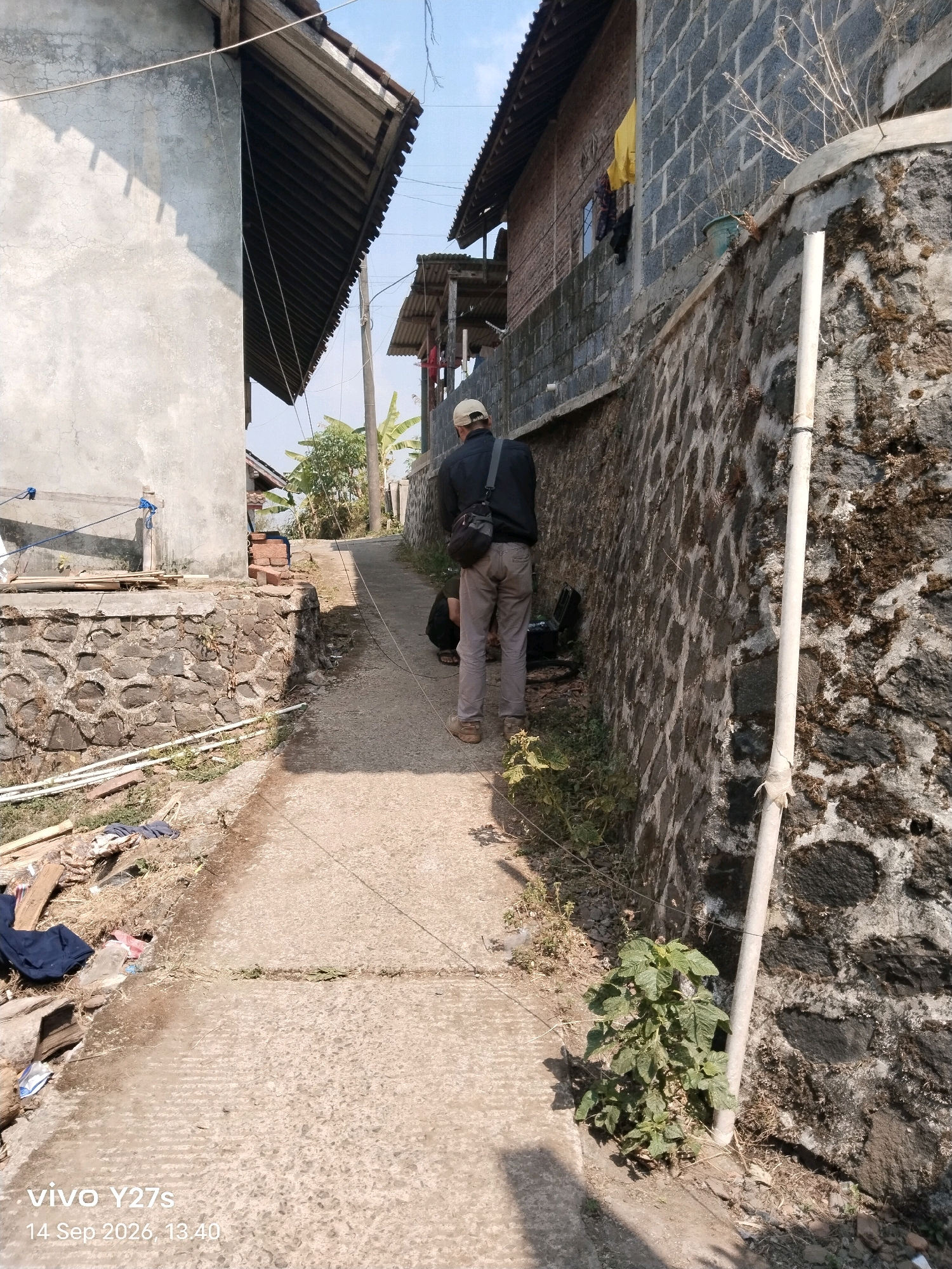 A narrow, paved alleyway winds uphill between two buildings. On the left, a light-colored, stucco-walled building with a corrugated metal roof has a low, stone-faced retaining wall at its base. Debris, including wood and fabric, is piled on the ground near the retaining wall. On the right, a building constructed of dark gray cinder blocks stands against a steep, stone-faced embankment. A white pipe runs vertically along the embankment wall. A man wearing a light-colored baseball cap, dark jacket, and light-colored pants is walking uphill in the alleyway, carrying a black bag over his shoulder. He is positioned in the center of the alley, facing away from the camera. Another person is partially visible, kneeling near the man on the right side of the alley. Banana trees and other vegetation are visible further up the alley. The sky is clear and blue. The image is dated "14 Sep 2026, 13:40" and has the watermark "vivo Y27s".
