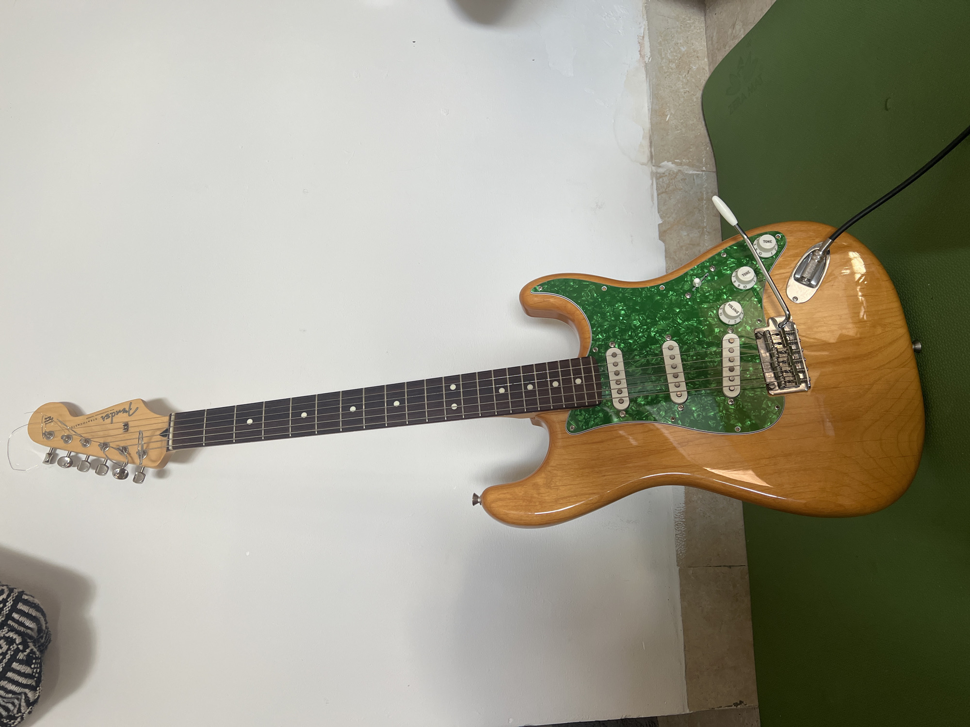 A Fender Stratocaster electric guitar with a natural wood finish and a green pearloid pickguard leans against a white wall. The guitar is positioned on a green yoga mat. The headstock, with its tuning pegs and "Fender Stratocaster" logo, is visible at the top. The body of the guitar features three single-coil pickups, volume and tone control knobs, and a tremolo bridge. A black cable is plugged into the guitar's output jack, extending out of frame. The background shows a plain white wall with some minor imperfections and discoloration towards the bottom. The floor appears to be tiled, with the green yoga mat placed on top. The scene is indoors, likely in a residential setting in Da Nang, Vietnam, as indicated by the presence of a yoga mat and the typical interior finishing. No people are present in the image.