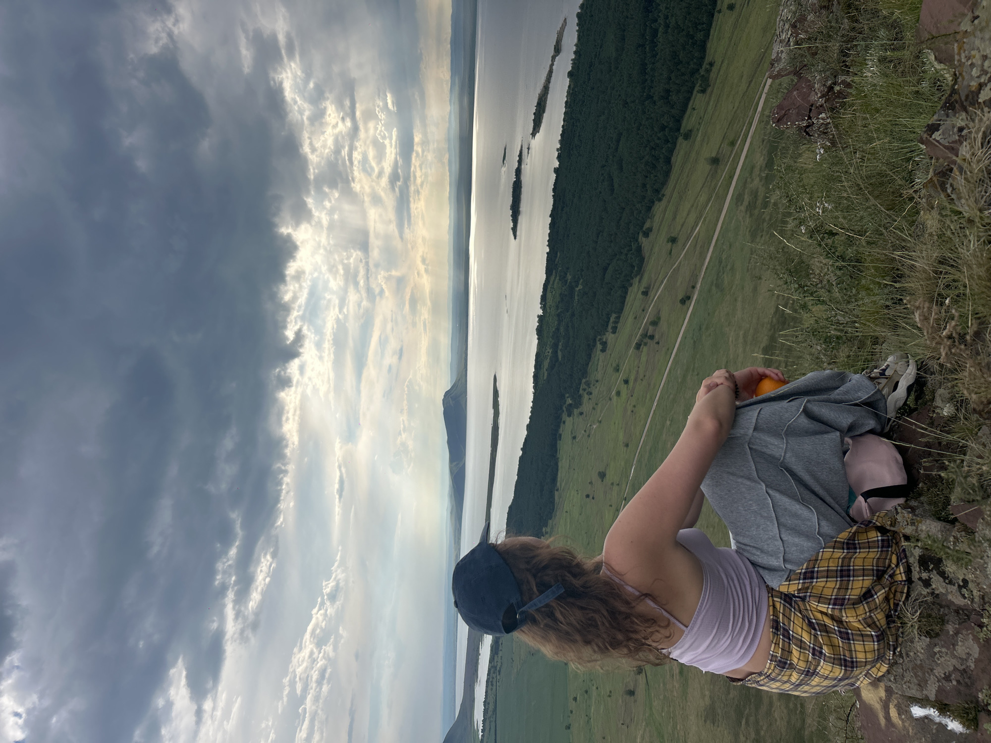 A young person sits on a rocky cliff overlooking a vast body of water and a treeline. They are wearing a light purple tank top, a yellow and black plaid skirt, and a dark blue baseball cap. They are holding an orange in their hands and appear to be wearing grey sweatpants and athletic shoes. The person is facing away from the camera, looking out at the expansive view.
The scene is set during the daytime, with a dramatic sky filled with both dark, brooding storm clouds and patches of bright sunlight breaking through. The light creates rays that illuminate parts of the clouds and the landscape below. The body of water is calm and reflects the sky, with several small, tree-covered islands dotting its surface. Beyond the water, a dense green forest stretches towards distant, hazy mountains. The foreground consists of rugged, rocky terrain covered in dry grass and some sparse vegetation. A dirt path is visible winding through the green fields on the lower slope of the landscape.
The overall mood is contemplative and serene, with a sense of awe inspired by the dramatic natural scenery. The contrasting elements of the stormy sky and the peaceful water suggest a moment of reflection amidst changing conditions. No text is visible in the image.