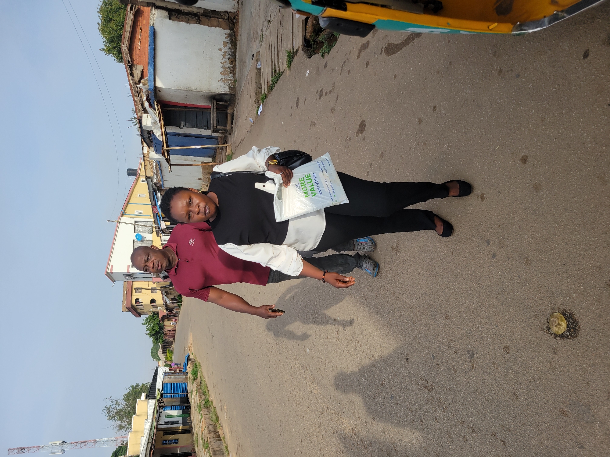 Two individuals stand on a paved road in what appears to be a residential or commercial area in Jos, Nigeria. The woman wears a black and white top and black pants, carrying a white plastic bag. The man wears a maroon shirt and jeans. The environment includes buildings of varied construction styles, from single-story structures with corrugated metal roofs to multi-story buildings. An antenna structure is visible in the distance. The road surface is visible, with the individuals standing on it. An object or spill is visible on the road's edge. A vehicle, possibly a taxi, is partially visible in the upper right corner.
