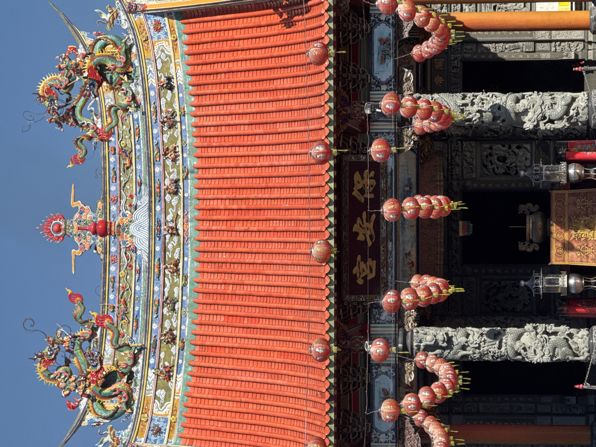 This is an upward-angled, outdoor shot of a traditional building, likely a temple, in Denpasar, Indonesia. The architecture is characterized by a multi-tiered, bright orange tiled roof. Ornate decorations, featuring colorful, sculpted dragons and other mythical creatures, adorn the roof's edges and corners. Strings of red lanterns hang from the building's eaves, suggesting a festive atmosphere.
The building's structure incorporates detailed carvings, particularly visible on the pillars. The sky is a clear blue, providing a stark contrast to the building's vibrant colors. The image is taken from below, emphasizing the height and grandeur of the temple. The image is rotated 90 degrees counter-clockwise.