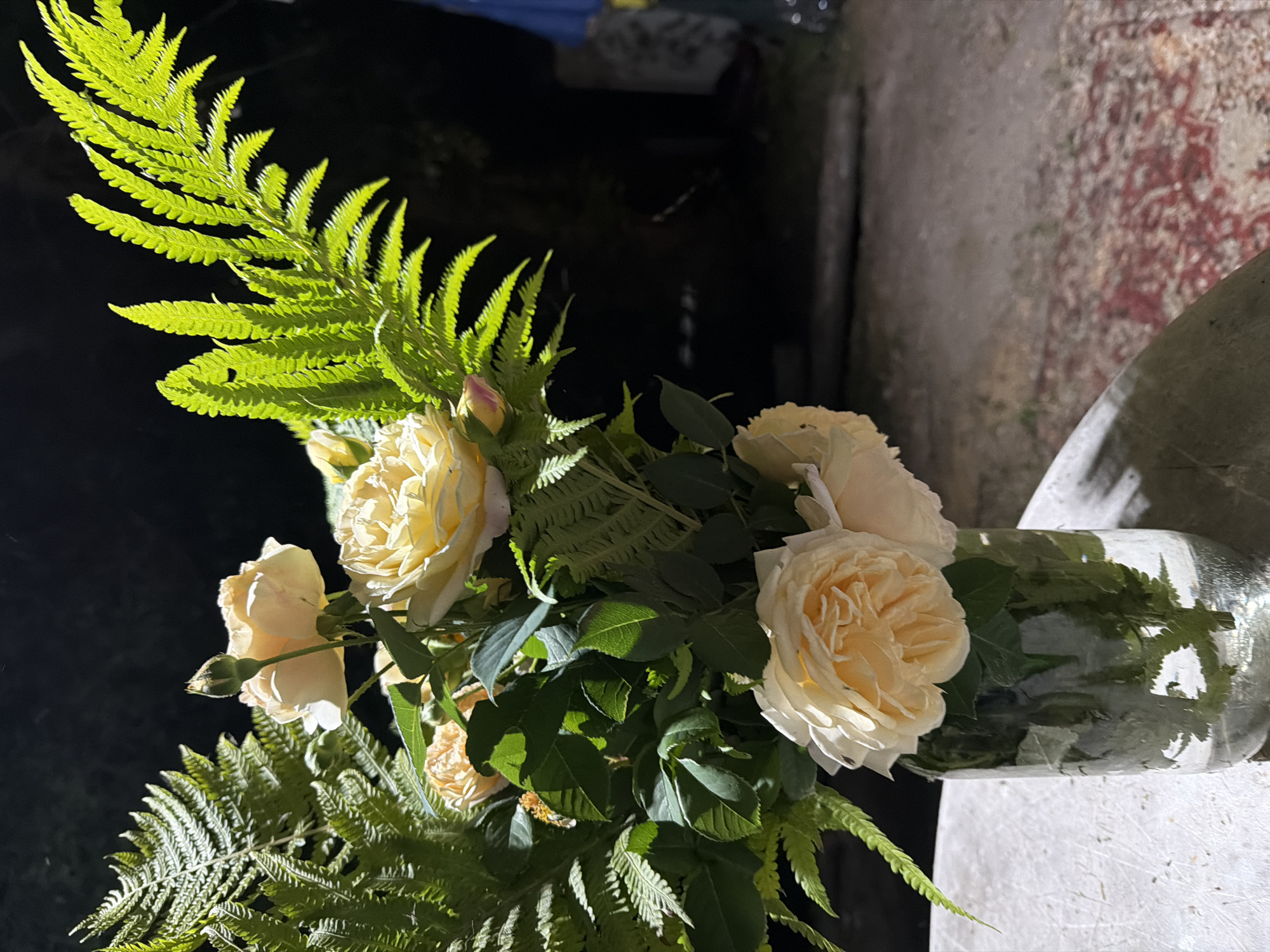 A vase filled with water and a bouquet of yellow roses and ferns sits on a textured surface that appears to be a table or stone. The bouquet is illuminated by a light source from the left, creating strong highlights and shadows on the flowers and foliage. The background is dark and out of focus, suggesting nighttime or a dimly lit environment. In the far right background, hints of a blue object and a blurry figure can be seen. The overall scene is naturalistic and captures a moment of floral arrangement in an outdoor setting.