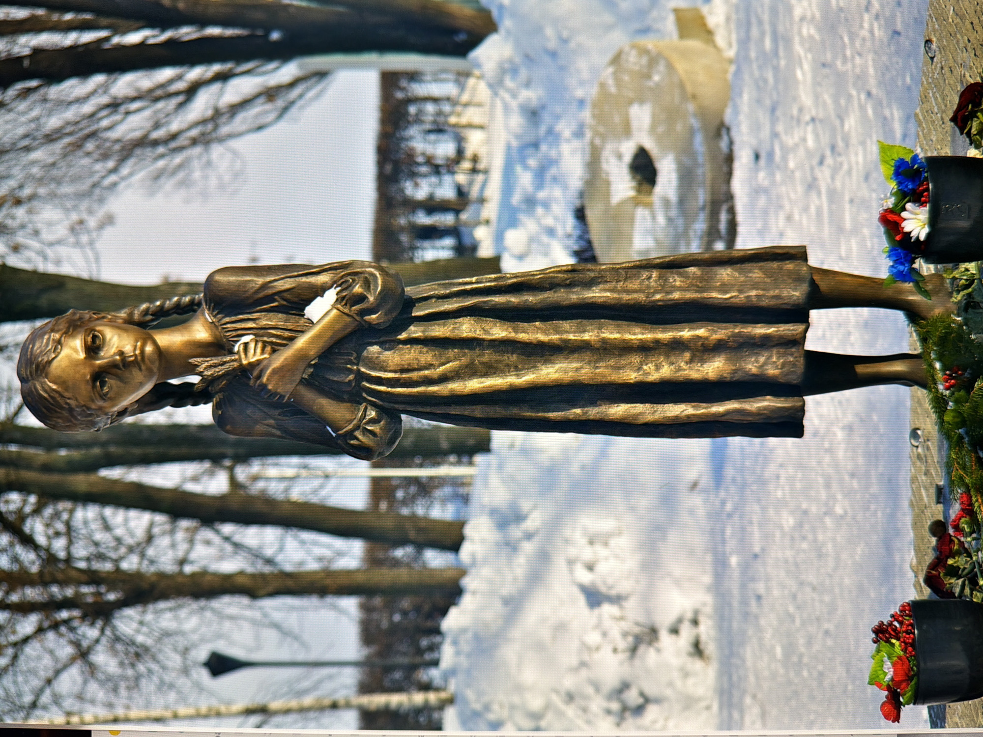 The image depicts a bronze statue of a young girl standing outdoors in a snowy environment. The girl is depicted with braided hair, wearing a long, pleated dress, and holding a bundle of wheat or stalks in her arms. She appears solemn and is looking forward. In the foreground, two black pots containing floral arrangements are placed. One arrangement features red flowers and berries, while the other has a mix of red, white, and blue flowers, along with some greenery. A circular stone structure with a dark opening in the center is visible in the background, partially covered by snow. The background also shows a line of bare trees and a lamppost. The overall scene suggests a memorial or a place of remembrance. The provided context indicates the location is in Kyiv, Ukraine.