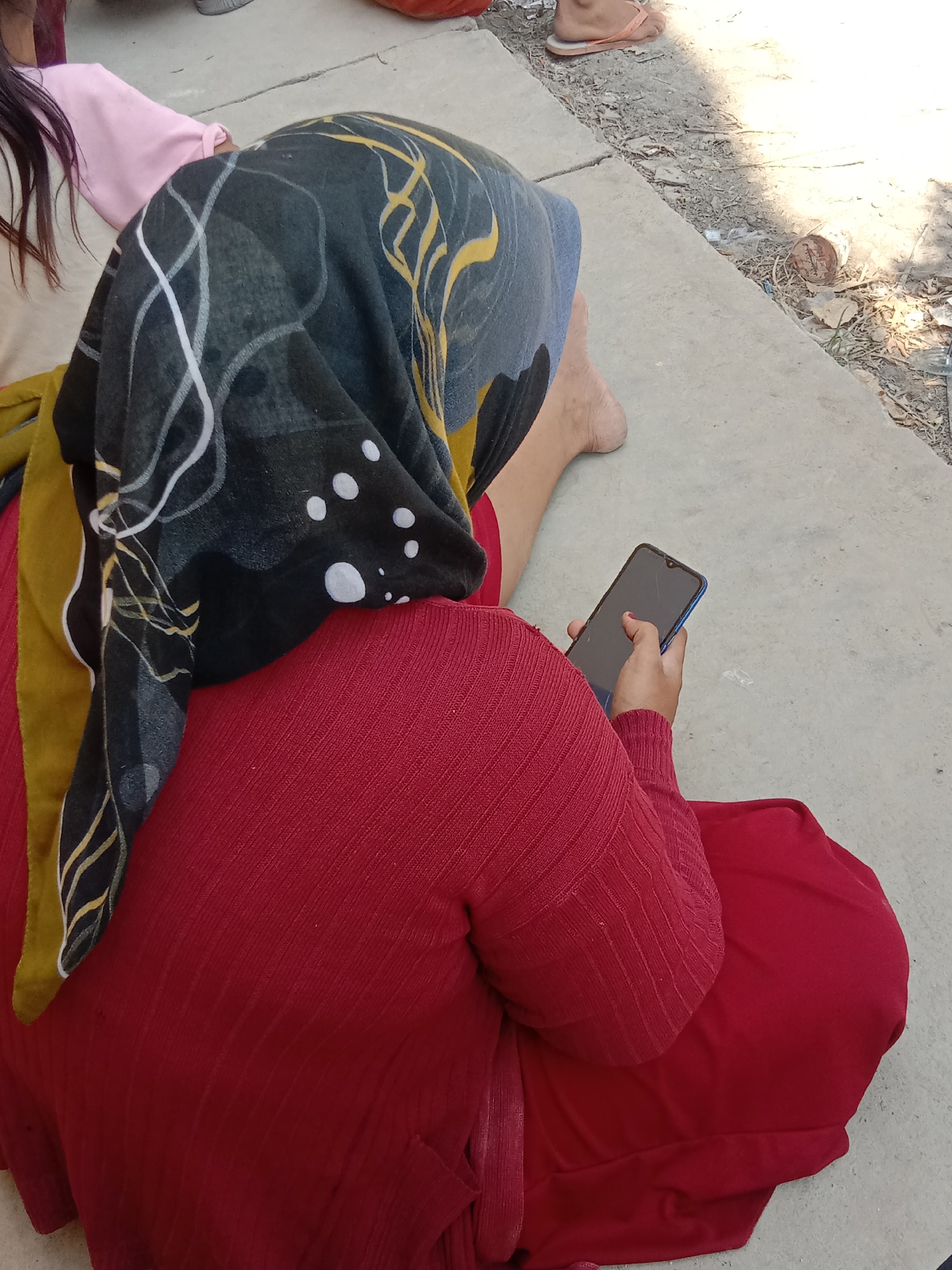 A person wearing a red top and a patterned headscarf sits on concrete, holding a blue smartphone. The person is viewed from behind. The background shows a dusty ground with some debris and another person's feet wearing flip-flops. The scene appears to be outdoors, possibly in a public area. The location in Kunden, Indonesia is not discernible from the image.