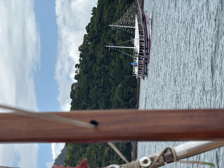 The image depicts a maritime scene in Angra dos Reis, Brazil, under a partly cloudy sky. In the foreground, a blurred reddish-brown wooden vertical beam, identified as part of a vessel's mast or rigging, occupies the left side of the frame, showing ropes and metal fixtures. A small red fabric detail is visible at the extreme upper left. In the midground, a white tour boat with red and dark blue trim, featuring two decks (one enclosed, one open) and two masts, is underway, moving from right to left across rippled grey-blue water. The masts are adorned with numerous small yellow triangular pennants. Multiple indistinct human figures, presumed to be passengers, are visible on both decks of the tour boat. The background features a densely forested, tropical green mountain or island coastline bordering the water. The sky above displays a mix of white cumulus clouds and segments of clear blue.