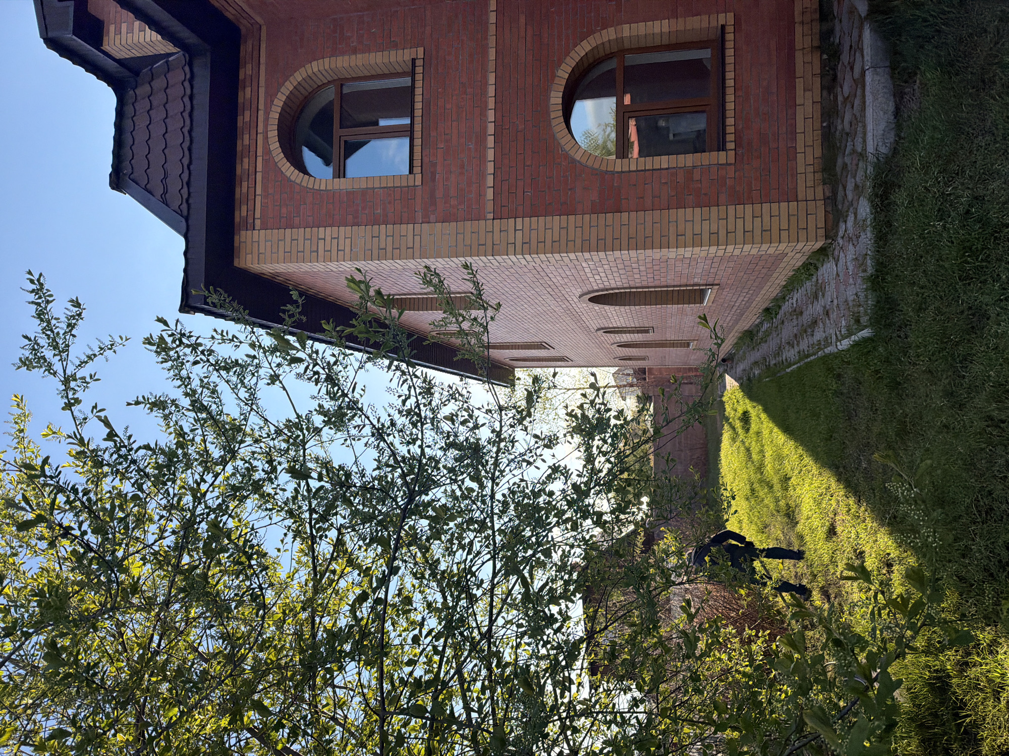 A brick building with distinctive round windows is viewed from a low angle, through a tree's branches. The building features contrasting red and beige brickwork. A narrow pathway runs along the side of the building. The presence of grass and greenery suggests a garden or yard surrounding the building. The sky is visible in the upper-left corner of the frame. The scene appears to be photographed outdoors, with natural light illuminating the building and surrounding landscape. The location is Krasnoyarsk, Russia.