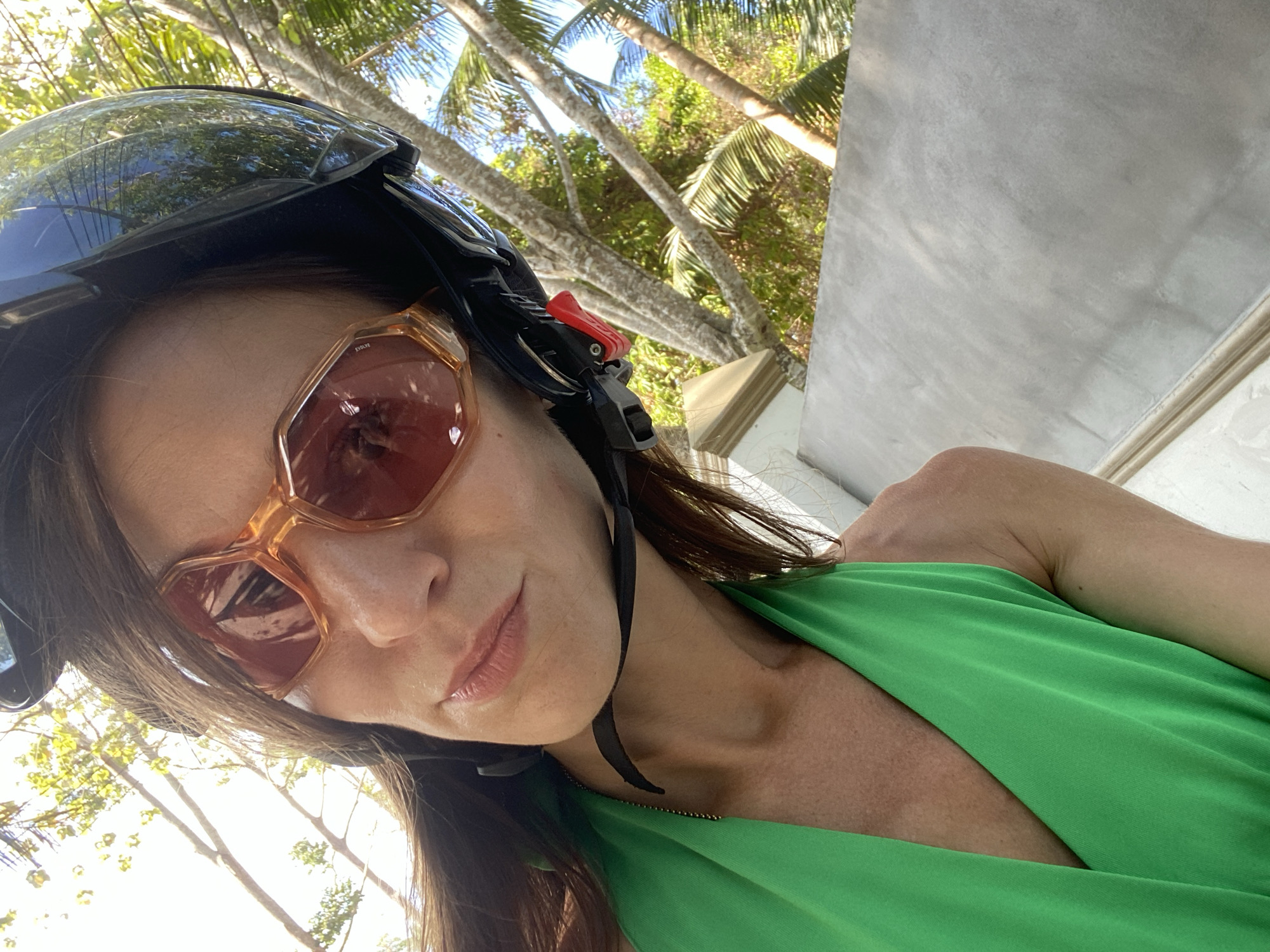 A selfie-style portrait captures a woman wearing a black motorcycle helmet with a clear visor and stylish hexagonal sunglasses with light brown frames. The text "ETOLIF" is faintly visible on the right temple of her sunglasses. She has long, dark brown hair and a neutral to slightly relaxed expression, with her lips slightly parted. She is dressed in a vibrant green halter-neck or wrap-style top, featuring a gold-colored zipper or trim near her neck.
The background depicts a sunny tropical environment, characterized by numerous tall palm trees with green fronds and other green foliage. To the right, a light gray, textured wall, possibly made of concrete or stucco, with decorative molding at its base, is visible. The bright sunlight and distinct shadows indicate it is daytime, likely midday or early afternoon, under clear weather conditions. The helmet and tropical scenery suggest she is either riding or about to ride a scooter or motorcycle, exploring a locale consistent with Moo 4, Thailand.