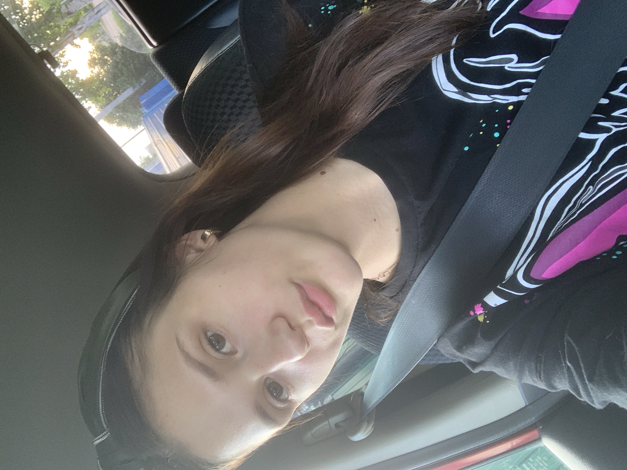 A young woman with long, dark brown hair is seated in the passenger seat of a car, facing forward, and looking at the camera. She is wearing a black t-shirt with a graphic design featuring pink and white shapes and colorful dots. A grey seatbelt is fastened across her chest. The interior of the car is visible, including the headrest of the seat behind her. Sunlight filters in through the side window, illuminating trees and a blue structure outside. The image appears to have been taken in Belogorsk, Russia, based on the provided context.