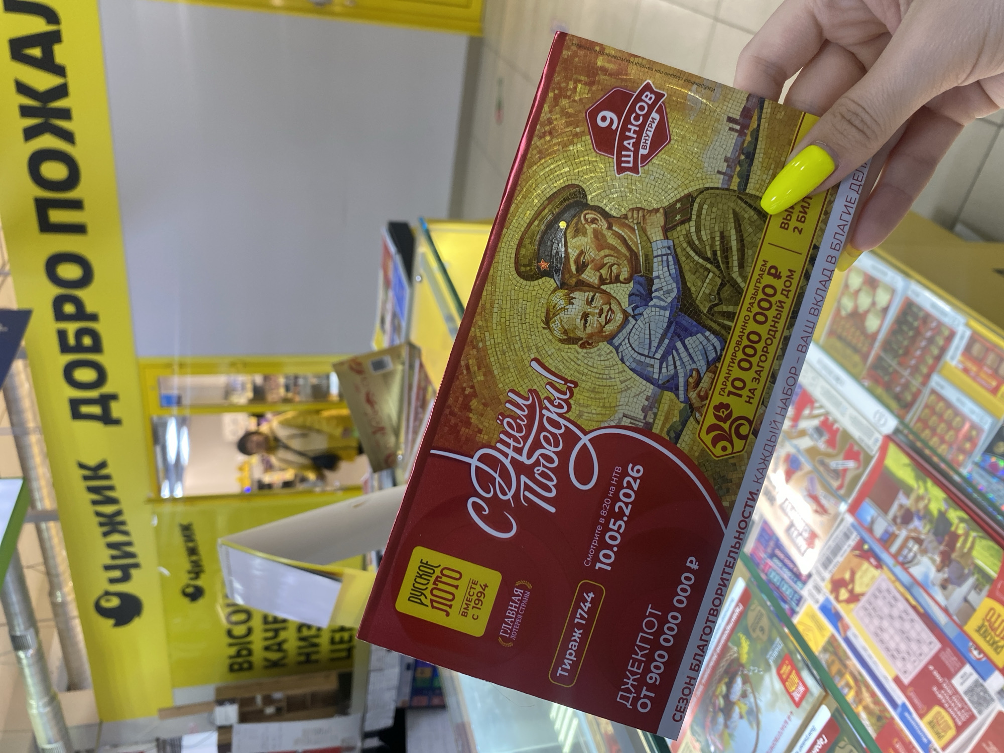 A hand with yellow fingernails holds a "Russian Lotto" lottery ticket in what appears to be a grocery store in the city of Chaikovsky, Russia. The ticket commemorates Victory Day ("День Победы!") and advertises a jackpot of 900,000,000 rubles. The drawing is scheduled for May 10, 2026. The ticket features an image of a soldier embracing a child. A sign for the "Чижик" (Chizhik) grocery chain is visible in the background. Lottery tickets are displayed on a shelf behind the hand holding the lottery ticket. The shelves are filled with various lottery tickets. A person is visible in the aisle of the store.
