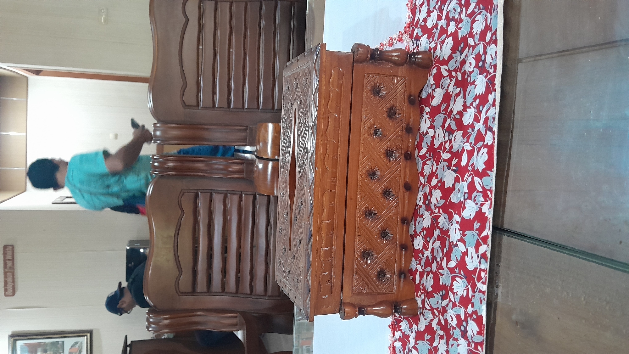 In a room possibly located in Blora, Indonesia, two men are present. One man, wearing a blue t-shirt, is facing away from the camera and appears to be holding a mobile phone. The other man, wearing a dark shirt and a cap, is seated and looking down. In the foreground, a carved wooden tissue box sits on a red and white floral fabric runner. Behind the tissue box are two wooden chairs with slatted backs. The overall environment suggests an indoor setting, likely a home or a common area, with wooden furniture and wall decorations.