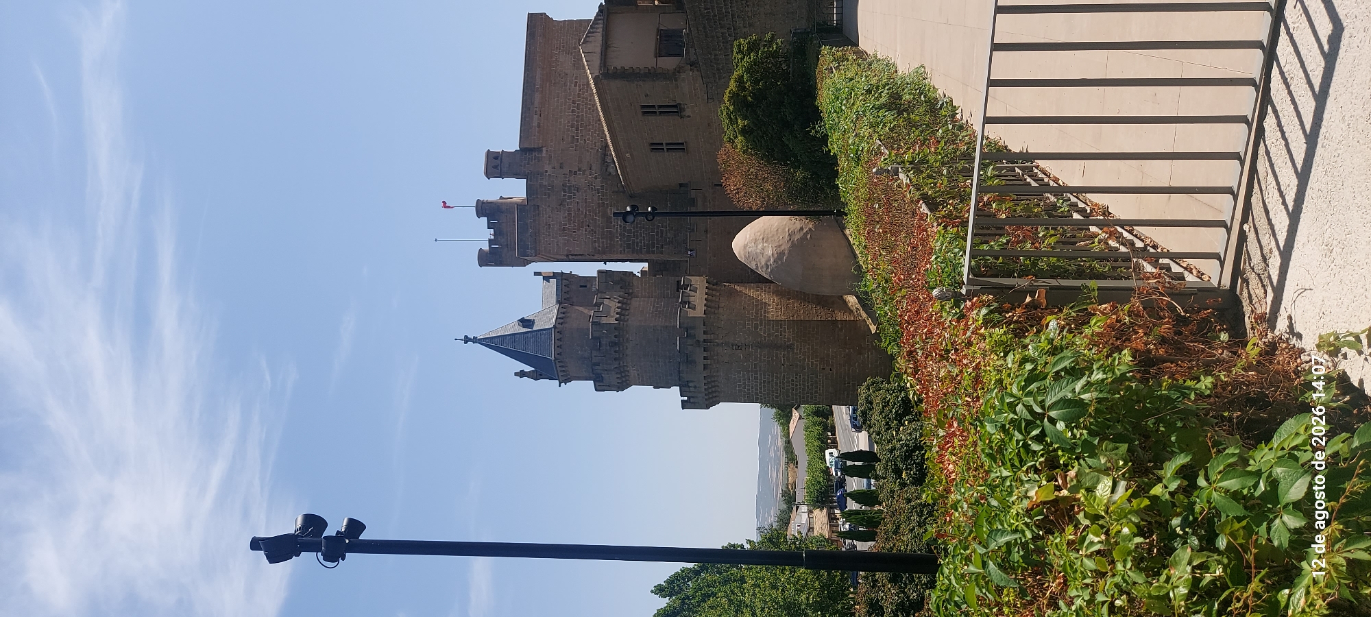 The image shows the Royal Palace of Olite, also known as the Palace of the Kings of Navarre, located in Olite, Spain. The palace is a medieval castle with multiple towers, including a prominent conical-roofed tower and a larger, square tower flying a red flag. In the foreground, a black lamppost with two lights stands tall against a blue sky with scattered white clouds. Lush green trees and colorful foliage are visible on the left and in the middle ground, with a road and some vehicles in the distance. A stone wall with battlements surrounds parts of the palace. A curved, egg-shaped stone structure is situated near the base of one of the towers. A metal railing separates a paved walkway from the vegetated area. The lower left corner of the image displays a timestamp: "12 de agosto de 2026 14:07".