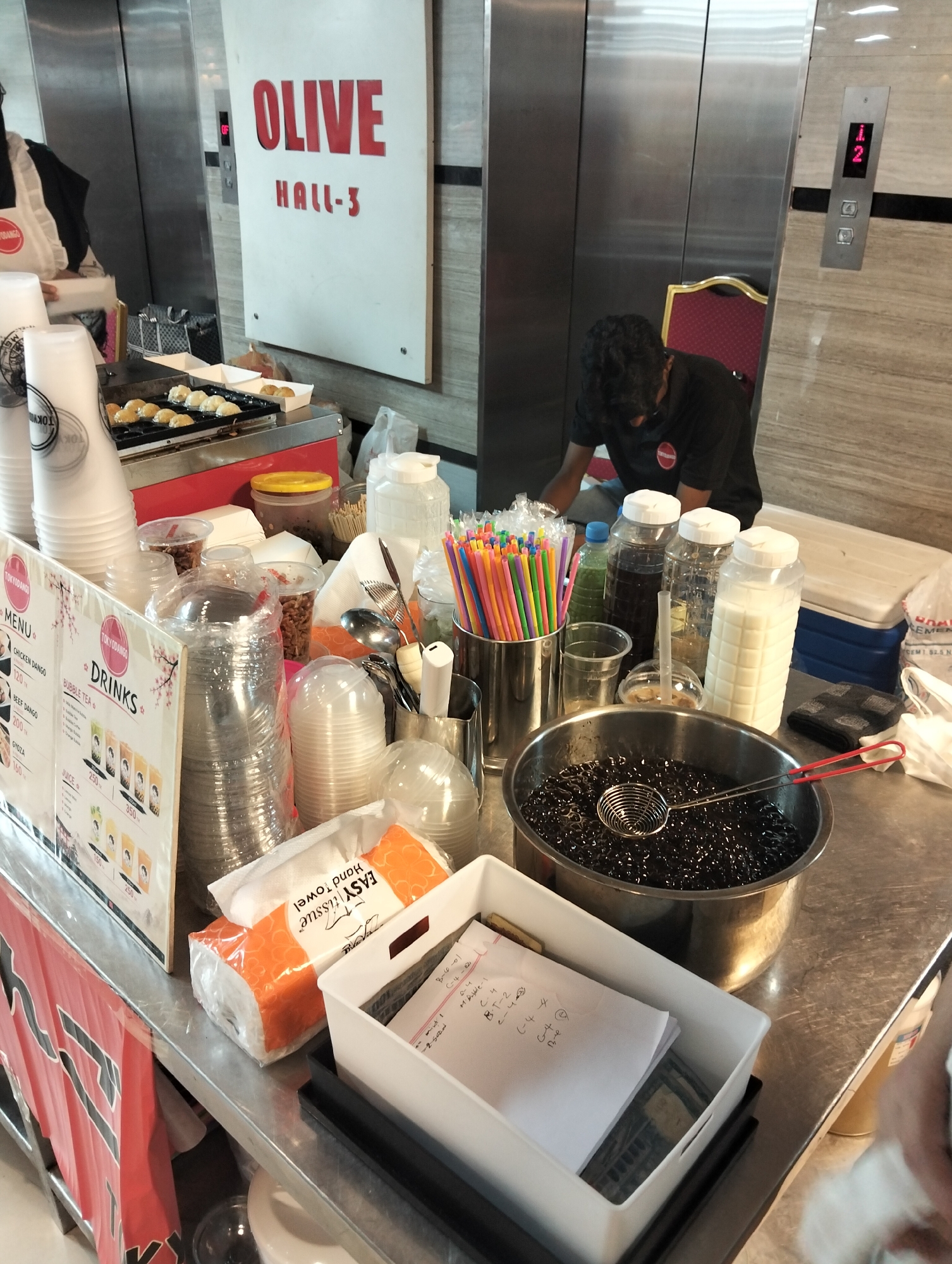 The image shows an indoor scene, likely a food stall or kiosk. In the background, an elevator door with illuminated numbers "1" and "2" is visible, along with a sign that reads "OLIVE HALL-3". A person wearing a black t-shirt is partially visible behind a counter, appearing to be working. The counter is laden with various items, including stacks of disposable cups, plastic lids, colorful drinking straws, large bottles containing dark liquid and white liquid, and a metal pot filled with black tapioca pearls. There are also packets of tissues, a white plastic container with some papers inside, and a grill with several small, round food items cooking on it. The overall environment appears to be a commercial space with a metallic, possibly stainless steel, countertop and walls.