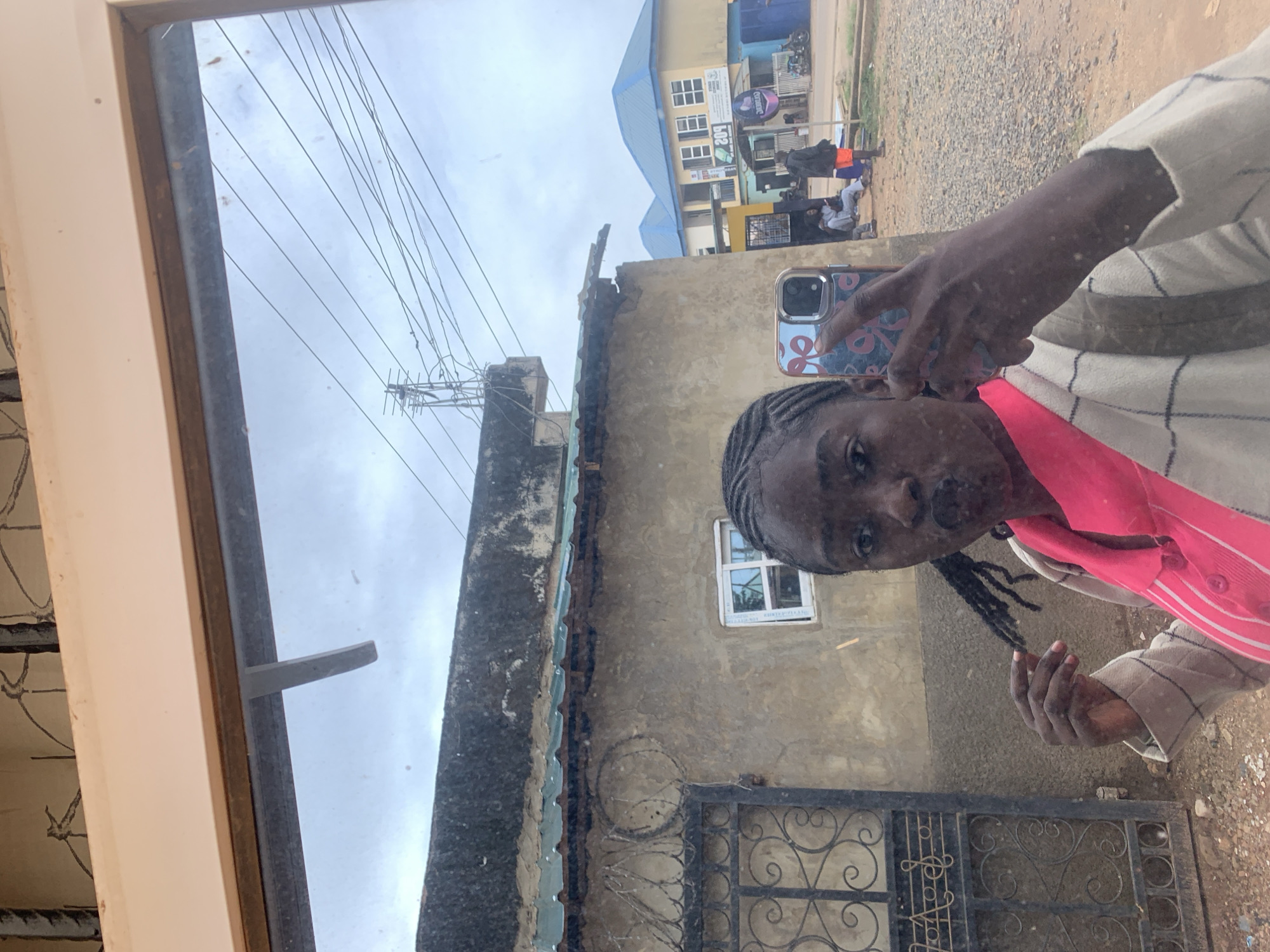 The image is a mirror or reflective window selfie of a dark-skinned female, taken in an outdoor urban setting in Jos, Nigeria. The female is positioned centrally, looking directly into the reflection. She has braided hair, part of which she holds in her left hand, and her lips are pursed in a pout. She wears a bright pink collared shirt beneath a light-colored, possibly off-white, jacket with a subtle dark plaid or grid pattern. Her right hand holds a smartphone, equipped with a patterned case featuring light blue and pink bow/ribbon designs, up to capture the image.
The reflection reveals a detailed street scene behind her. In the immediate background, a weathered, light brown/tan building with a rough textured facade and a dark, possibly concrete, flat roofline is visible. Below the roof, a green corrugated metal awning or overhang extends. To the left of this building is an ornate black wrought-iron gate with scrollwork. A small, white-framed window is set into the building's wall. The ground in the foreground of the reflection is unpaved, appearing as dirt and gravel.
Overhead, numerous black electrical power lines crisscross against an overcast, grey sky. A utility pole with multiple wires and a small antenna-like structure is present. Further into the background, a multi-story yellow building with a prominent bright blue corrugated metal roof stands. Commercial signs are visible on its facade, one partially legible as "209". Another similar blue-roofed building is partially visible behind it. Several individuals are gathered on the street or sidewalk near the yellow building, including one person in an orange top and dark shorts standing prominently, and others seated or standing further back. A motorcycle is parked on the far right of the reflection. The entire reflected scene is framed by a simple, off-white or light-colored window or mirror frame along the top and sides.