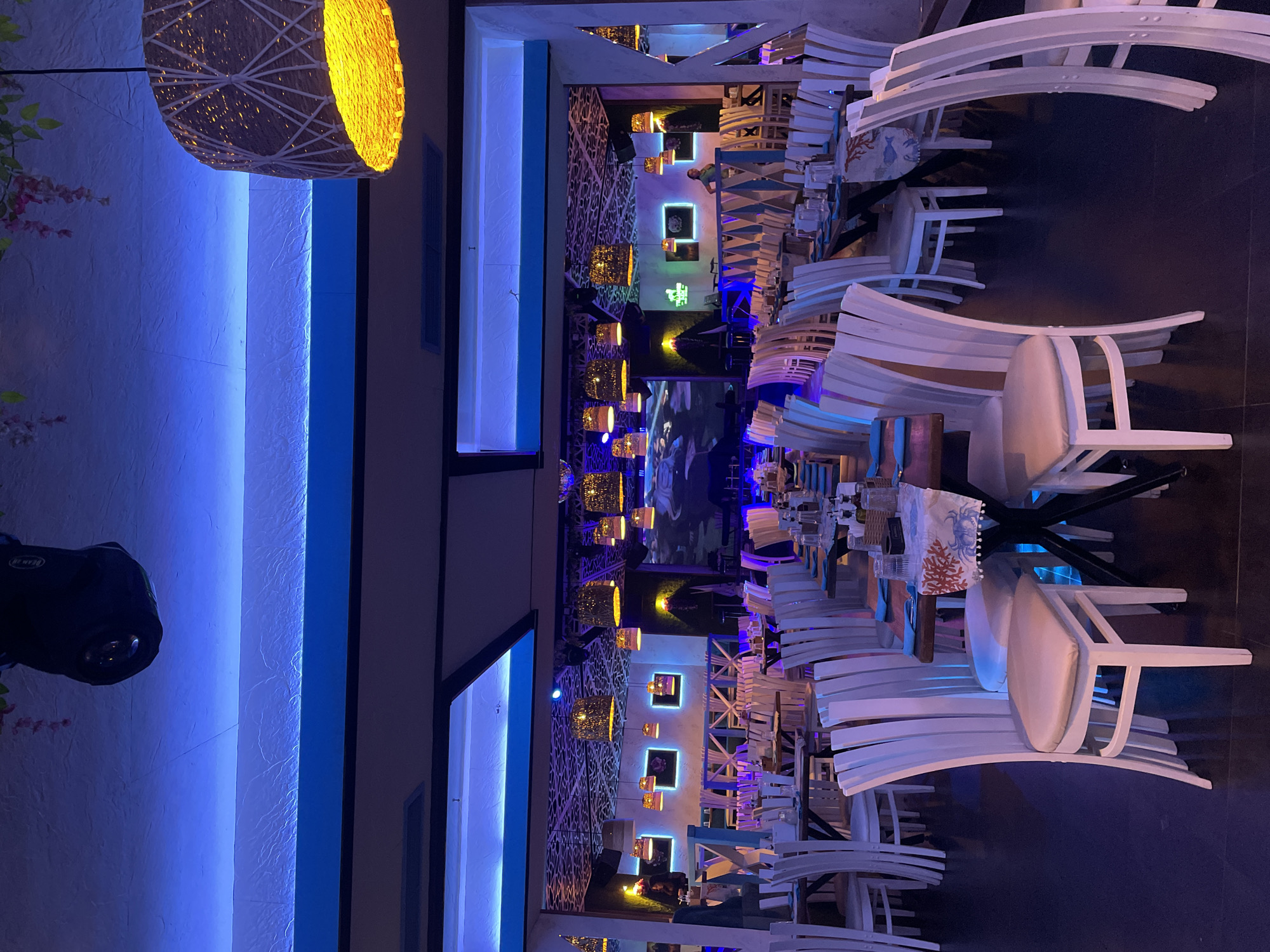 The image depicts the interior of a restaurant in Plovdiv, Bulgaria, characterized by its modern design and distinctive lighting. The foreground features several white chairs with curved backs arranged around dark tables. The restaurant's interior is decorated with blue LED lighting strips on the walls and hanging wicker pendant lights. Further into the restaurant, there is a decorative wall with geometric patterns and more pendant lights. Portraits with lighting adorn a wall. A large screen or mural is visible in the middle of the room. There are no people visible in the immediate foreground. A black lighting fixture is fixed to the ceiling. The floor appears to be a dark color, contrasting with the white furniture.