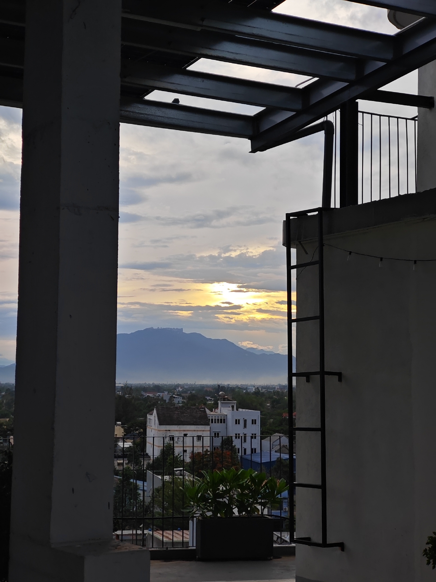 The image captures a panoramic view of Đà Nẵng, Vietnam, from an elevated vantage point, likely a balcony or rooftop. The foreground is dominated by a concrete pillar on the left and a white building with a metal ladder and string lights on the right, creating a framed perspective of the cityscape.
The midground reveals a cluster of buildings, including a white structure with multiple windows and a dark blue roof, nestled amongst lush green trees. In the distance, a majestic mountain range dominates the horizon, bathed in the warm, golden light of the setting sun. The sky above is a canvas of soft, diffused clouds, with the sun peeking through, casting a gentle glow over the entire scene. The overall ambiance is serene and tranquil, evoking a sense of peace and contemplation.