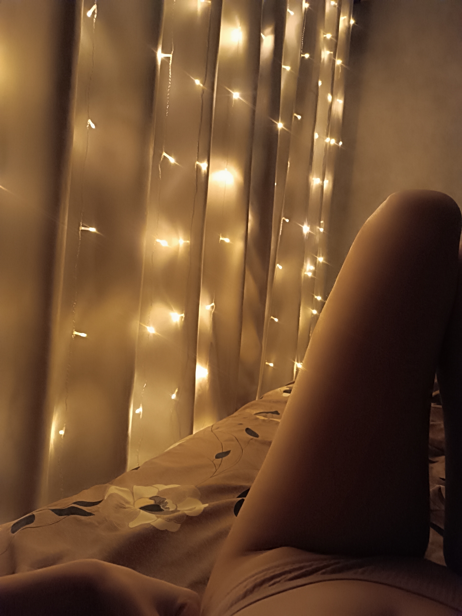 A dimly lit indoor scene shows a person lying on a bed with a patterned duvet cover. Strings of small, warm-toned lights are draped vertically in the background, resembling a curtain. The person's leg is the prominent subject in the foreground, suggesting a relaxed or resting posture. The environment is suggestive of a bedroom. The city of Unknown, Unknown, cannot be determined from the image.