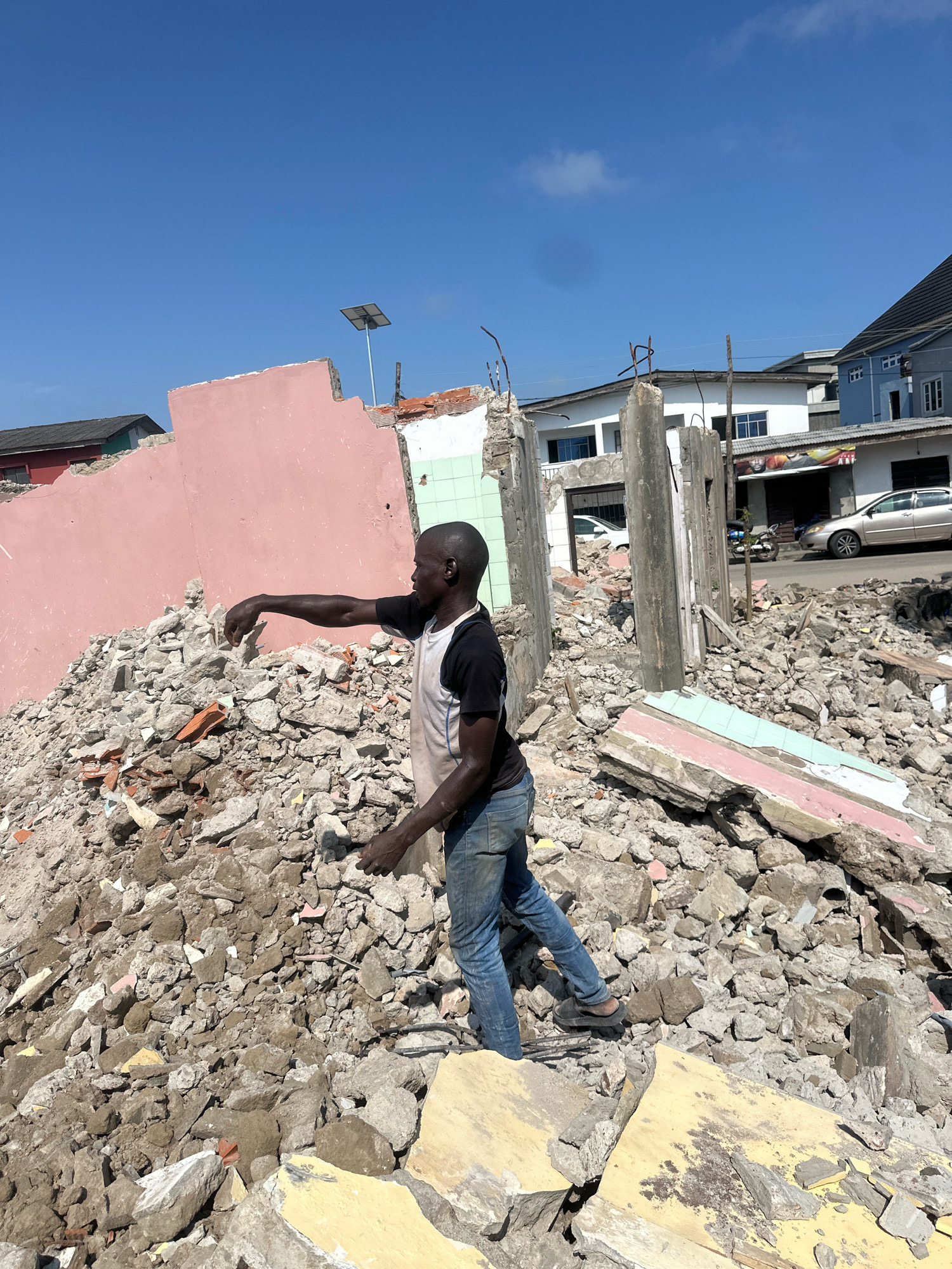 A man wearing a black and white t-shirt and blue jeans stands on a pile of rubble, gesturing with his right hand towards the debris. The rubble consists of broken concrete, bricks, and other building materials. Behind him, a pink wall partially remains from a destroyed building, with exposed rebar protruding from concrete structures. In the background, several buildings are visible, some appearing intact and residential, while others show signs of damage or are under construction. The sky is clear and blue. The scene appears to be in Badagry, Nigeria, likely a demolition or disaster site.
