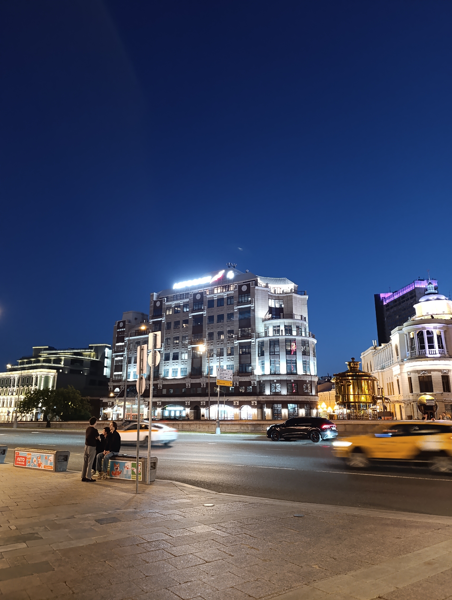 This night-time urban street scene in Moscow, Russia, depicts the area around Okhotny Ryad, Mokhovaya Street, and Teatralny Proyezd, viewed from a slightly elevated pedestrian area.
In the foreground, a wide, paved pedestrian sidewalk occupies the lower portion of the frame. Three individuals are gathered on the left side of the sidewalk: two are seated on a dark-colored public bench with a graphical advertisement featuring a squirrel and the Russian text "ЛЕТО В РОССИИ" (Summer in Russia), while a third person stands facing them. Another identical bench is partially visible to the right.
The midground is dominated by a multi-lane asphalt road. Multiple vehicles are in motion, depicted with motion blur: a bright yellow taxi moves right in the closest lane, a dark-colored SUV (possibly an Audi) follows in the lane to its left, and a white car is visible further left. Several street signs on poles are positioned between the sidewalk and the road. On the far side of the road, a large, ornate golden structure, resembling a traditional Russian samovar or kettle, stands prominently illuminated between buildings.
The background is filled with brightly lit architectural structures under a deep indigo-blue sky. The central and most prominent building is a multi-story edifice with a light-colored facade, numerous windows, and a distinctive curved section on its upper floors. A prominent illuminated sign, which appears to read "HOTEL NATIONAL," is visible on its rooftop. To the immediate right of this building, another elaborate structure features a distinctive domed cupola and classical columns. This is the Metropol Hotel. Further right, a taller, darker building with purple illumination near its top is partially visible. To the left of the central Hotel National, a classical-style building with white columns and bright lighting is seen, partially obscured by trees. Behind this, another modern, tiered building with white illumination extends upwards. A subtle crescent moon is visible in the sky above the central Hotel National. The overall lighting suggests artificial illumination from streetlights and building facades.