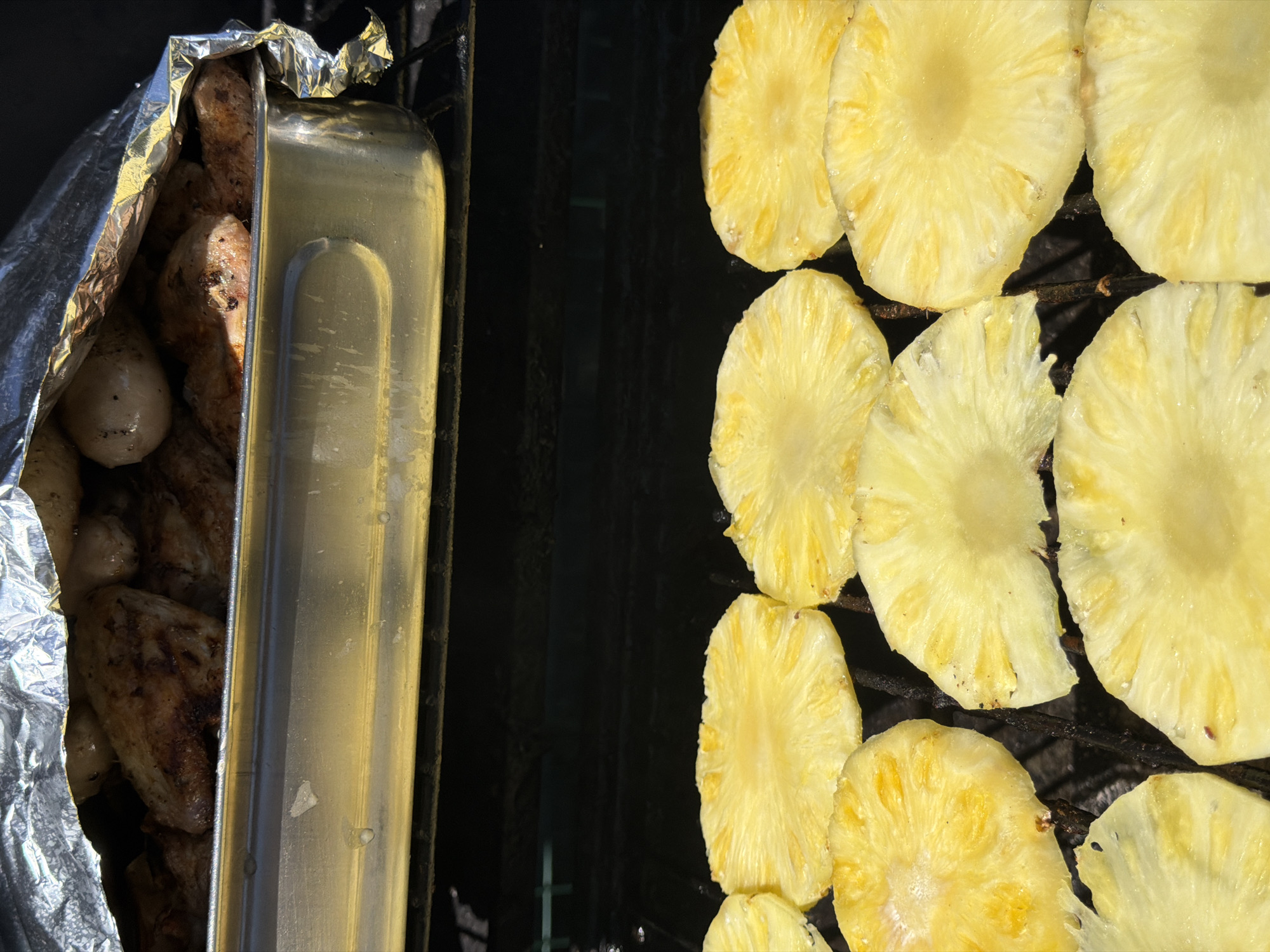 This image presents a vibrant, high-angle view of an outdoor grilling scene, likely taking place in Esch-sur-Alzette, Luxembourg. The frame is distinctly divided into two halves, showcasing different cooking methods.
On the left, a rectangular metal pan, possibly a disposable aluminum tray, is seen nestled within crumpled silver aluminum foil, suggesting an attempt to keep food warm or contain it while grilling. Inside this foil-wrapped container are several pieces of cooked meat, likely chicken or pork, displaying a golden-brown, seared texture with some visible seasoning and charring.
The right side of the image is dominated by numerous circular and semi-circular slices of fresh, bright yellow pineapple, arranged directly on a dark, sturdy metal grill grate. The pineapple slices show the characteristic fibrous pattern of the fruit, with some appearing slightly softened and lightly caramelized from the heat, indicating they are actively being grilled.
The scene is brightly lit by strong, direct sunlight, creating sharp contrasts and highlights on the reflective foil and the juicy pineapple, while casting shadows under the grill grates. The overall impression is one of preparing a tropical-themed barbecue meal during a clear daytime. There are no people visible, and no text is present in the image.
