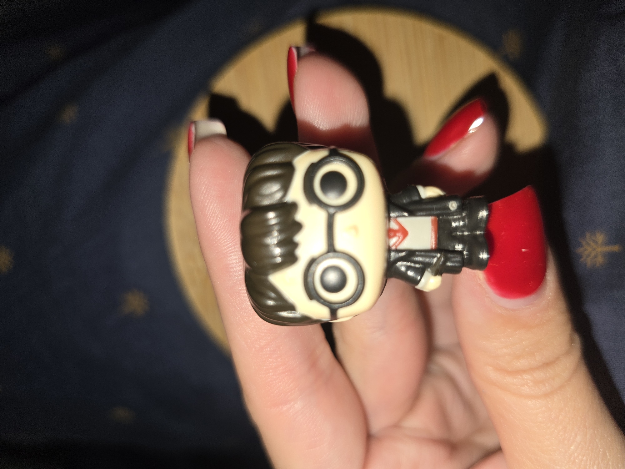 The image shows a close-up of a person's hand holding a Funko Pop figure. The figure is stylized with a large head and big black eyes with white circles. It has dark brown hair and is wearing a black outfit with red accents. The hand holding the figure has bright red fingernail polish. The background is dark blue fabric with a subtle pattern of yellow stars. A blurred wooden surface is visible behind the figure. The lighting suggests artificial light, possibly from a lamp or overhead fixture, casting shadows. The setting appears to be indoors, with no specific location cues other than the user-provided context of Petropavlivska Borshchahivka, Ukraine. No text is visible in the image. The activity appears to be someone displaying or playing with the collectible figure. The focus is on the hand and the figure, with the background out of focus.