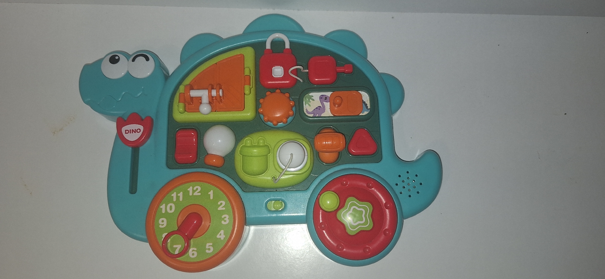 This is a top-down, close-up studio photograph of a turquoise plastic dinosaur-shaped baby activity toy placed on a white, neutral surface. The toy is oriented with its head to the right and its tail to the left. The toy has various interactive elements, including a clock with an orange face and red hands, a green rotating dial with star cutouts, several colorful buttons and levers in red, orange, and green, and a white ball connected to a string. The toy is illuminated by artificial light, casting subtle shadows. There are no people present, and the environment is a plain, unadorned surface, suggesting a product or commercial photography setup. The location of Bila Tserkva, Ukraine is not discernible from the image.