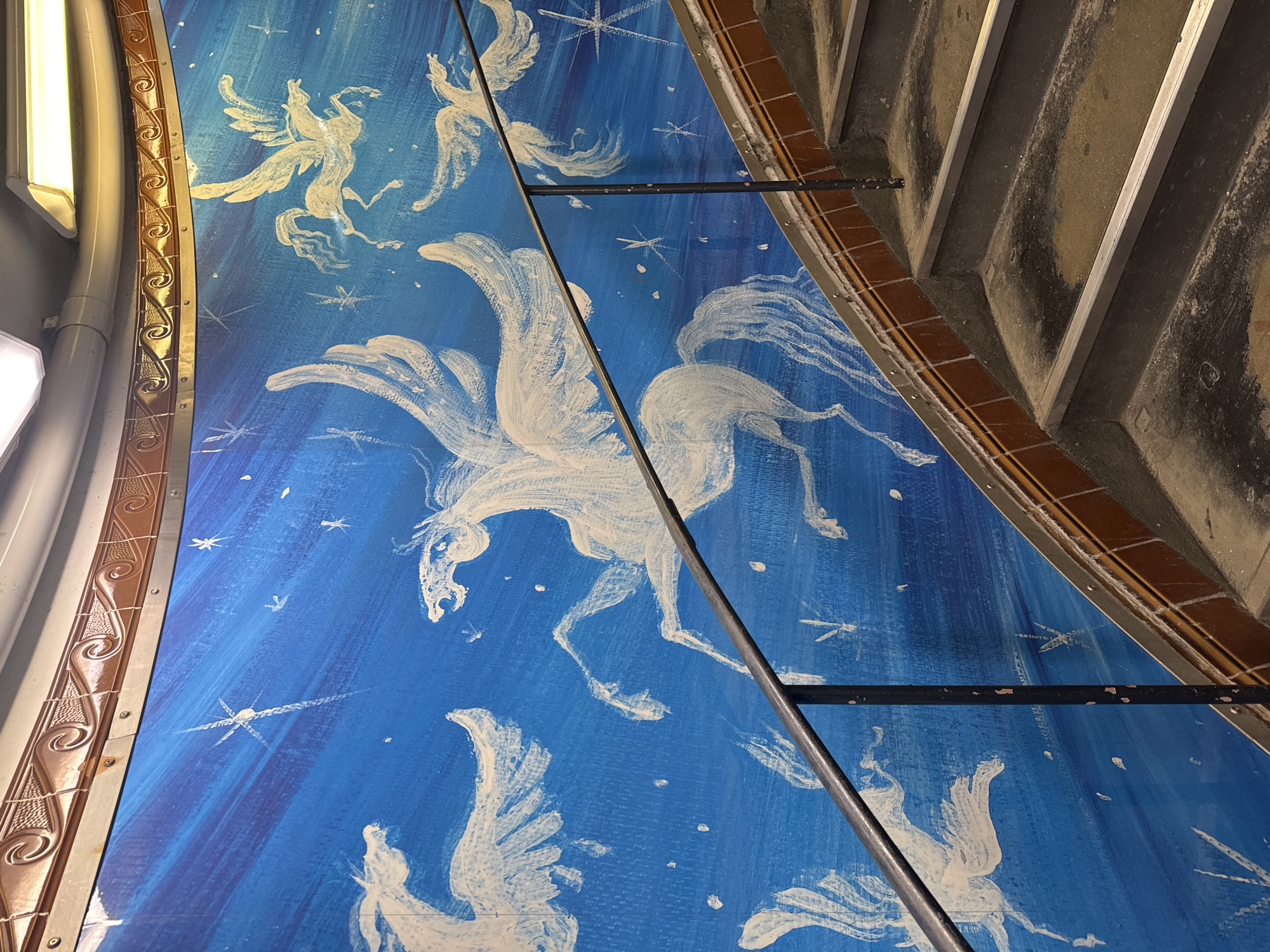 This image shows a section of the ceiling mural in the 191st Street 1 train station in New York City, featuring white Pegasus figures and stars painted on a blue background. Adjacent to the mural are architectural elements, including a patterned metallic trim along one side and a set of concrete stairs along another side. Metal support beams run across the mural's surface. A fluorescent light fixture is visible near the trim.