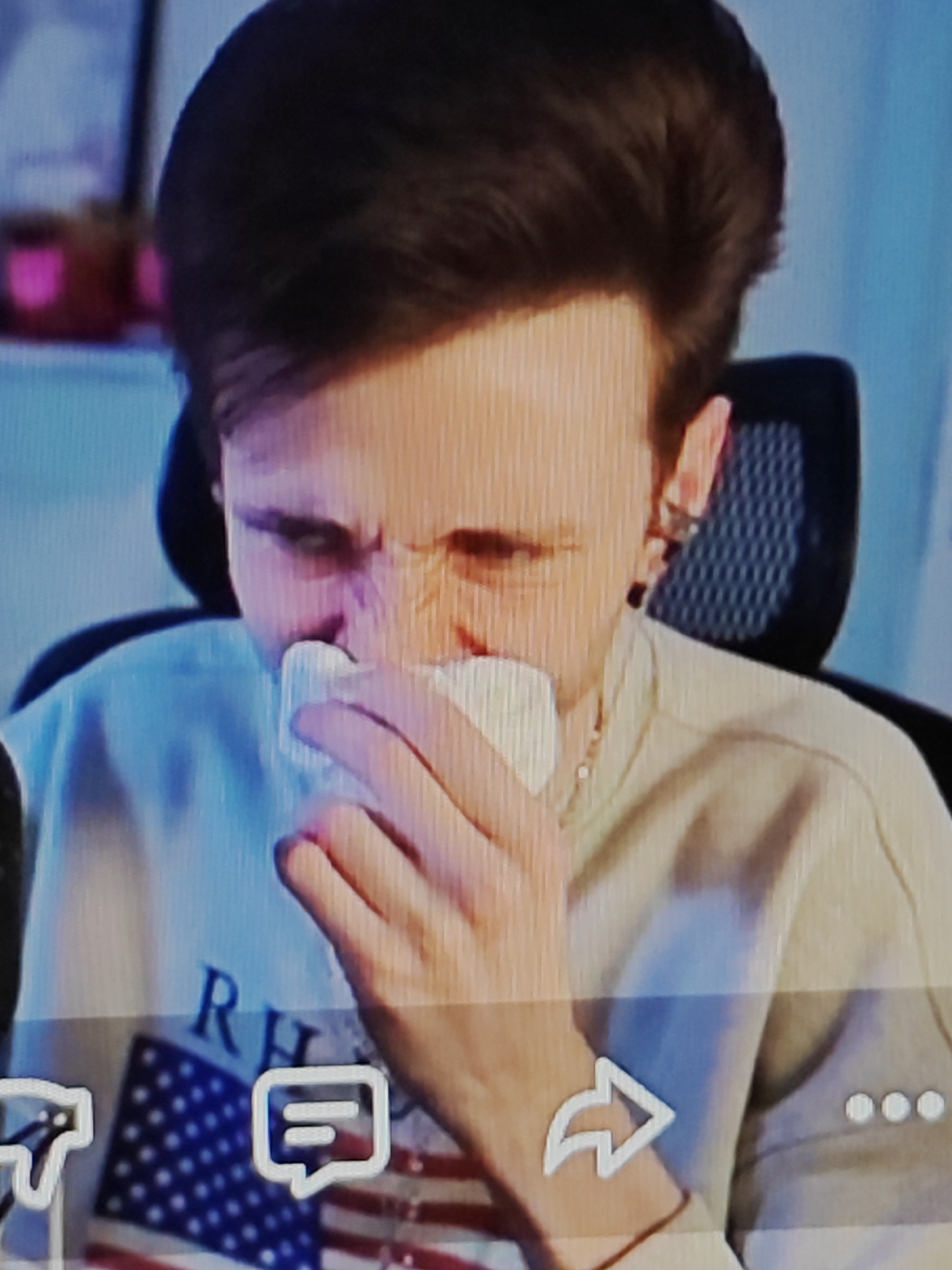 The image is a medium close-up shot of a person with brown hair holding a white tissue to their nose. The person is wearing a light-colored sweatshirt with an American flag graphic on the front, featuring the letters "RH" above it. The background is blurred, showing a blue wall and some pink objects on a shelf. There are also on-screen icons for a thumbs down, a chat bubble, a share arrow, and three dots, suggesting this is a screenshot from a video or online platform. The lighting appears to be artificial, casting a slight orange hue on the person's face. The scene is indoors, with the person seated in what appears to be an office chair. There is no indication of the location within a city or any specific geographical context.