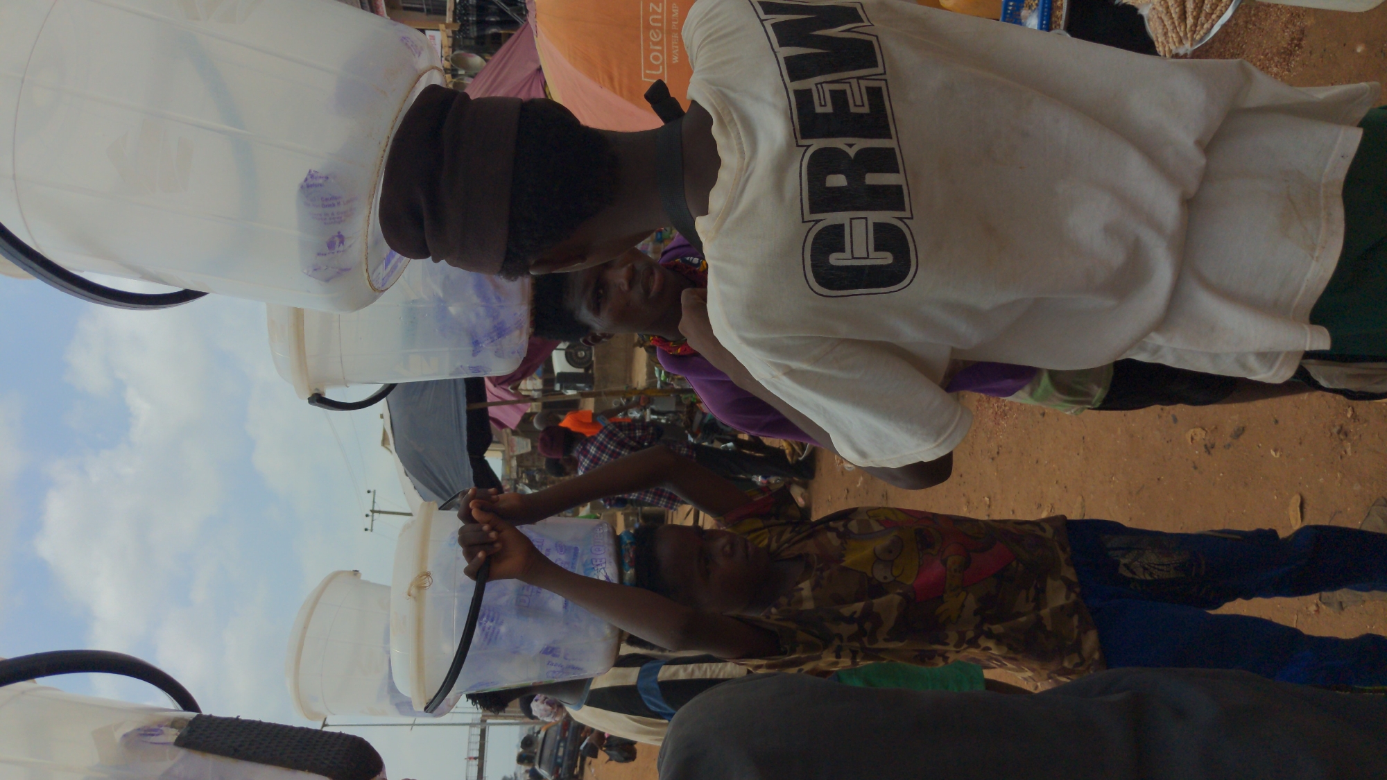 A street scene in Ado, Nigeria, shows several people transporting large white plastic buckets, presumably containing water, on their heads. The individuals are African, and one wears a white t-shirt with "CREW" printed on the back. Another person is carrying a bucket by its handle, with raised arms. Vehicles are visible in the background, along with utility poles and other pedestrians. The ground is dirt.