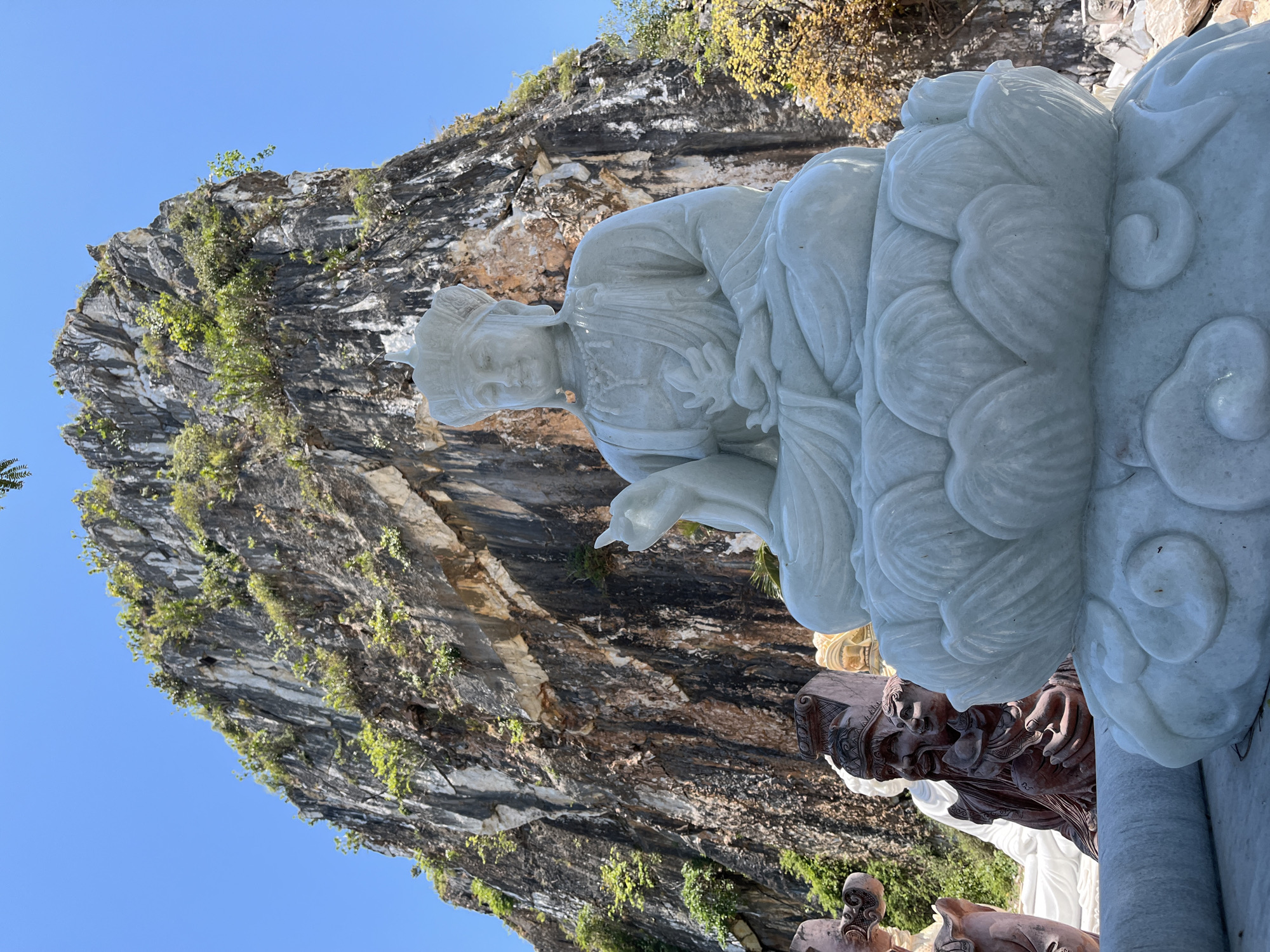 A medium shot shows a large white marble statue of a seated figure at the Marble Mountains in Đà Nẵng, Vietnam, against a rocky outcropping. The statue is seated on a stylized lotus platform. Other statues of various colors are visible in the lower foreground. The sky is blue and clear in the background.