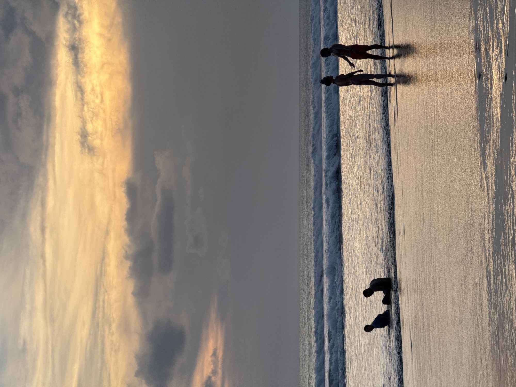The image depicts a sunset scene on Kuta Beach, Kuta, Bali, Indonesia. Two people are standing at the water's edge, and two people are crouching in the water. The sky shows golden clouds.