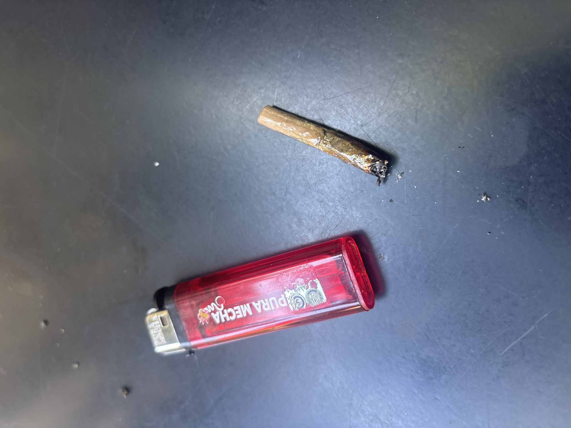 The image shows a partially smoked marijuana cigarette and a red "Pura Mecha" brand lighter on a dark, possibly metallic, surface. The cigarette is burnt at one end with visible ash. The lighter is a standard rectangular shape with a silver striking mechanism. The background is a dark, slightly reflective surface with some scattered debris. Without additional information, determining the specific location in Buenos Aires is impossible.