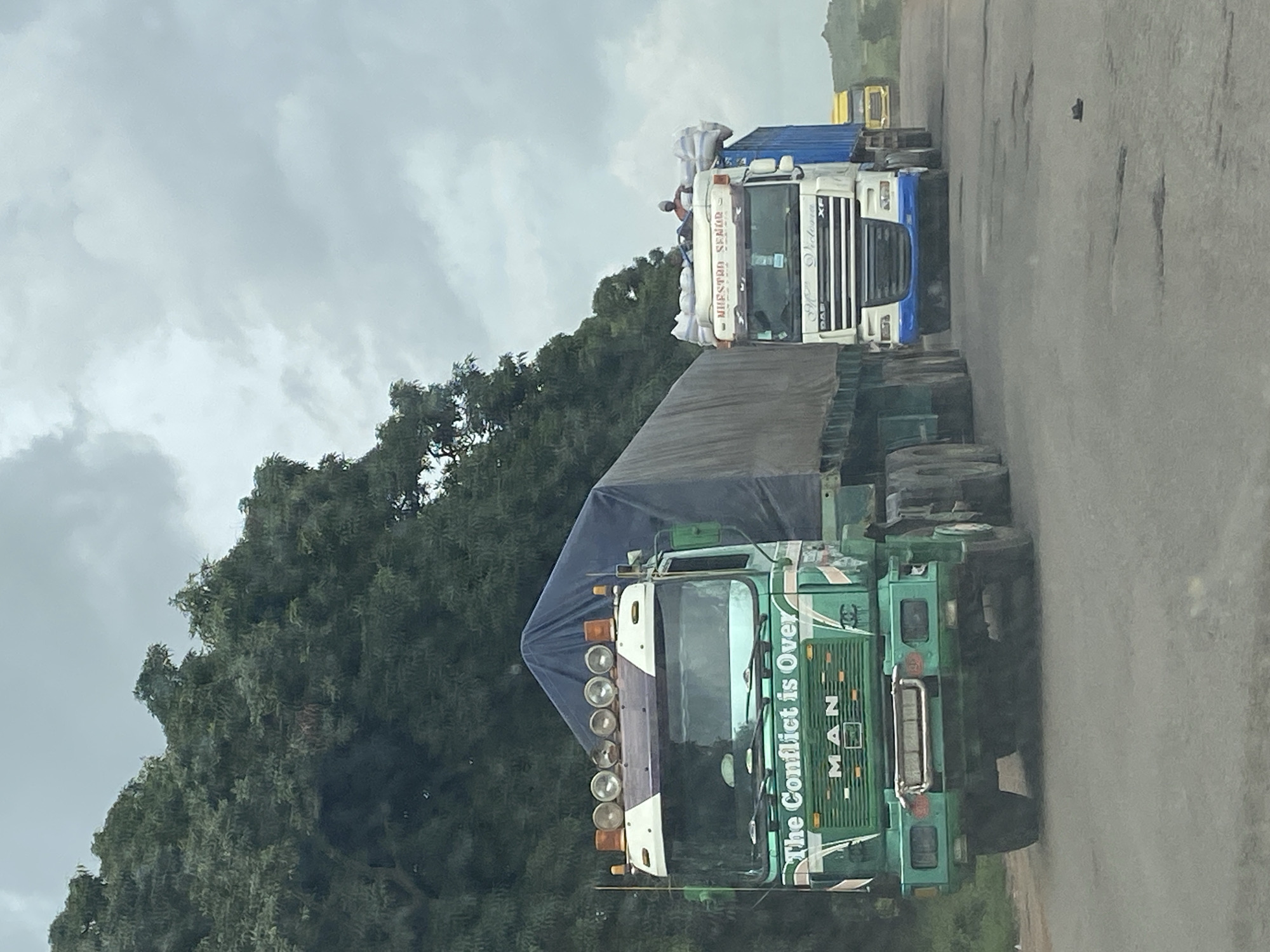 Two large trucks are seen on a paved road, with a line of trees in the background. The first truck, in the foreground and to the left, is green and has the words "The Conflict is Over" printed on its side. The second truck, to the right, is white and has a blue trailer. The sky is overcast. A person is visible on top of the second truck, near some bagged cargo. The road surface shows signs of wear and tear, with visible potholes. The image appears to be taken from another moving vehicle. The location is Kuru, Nigeria.