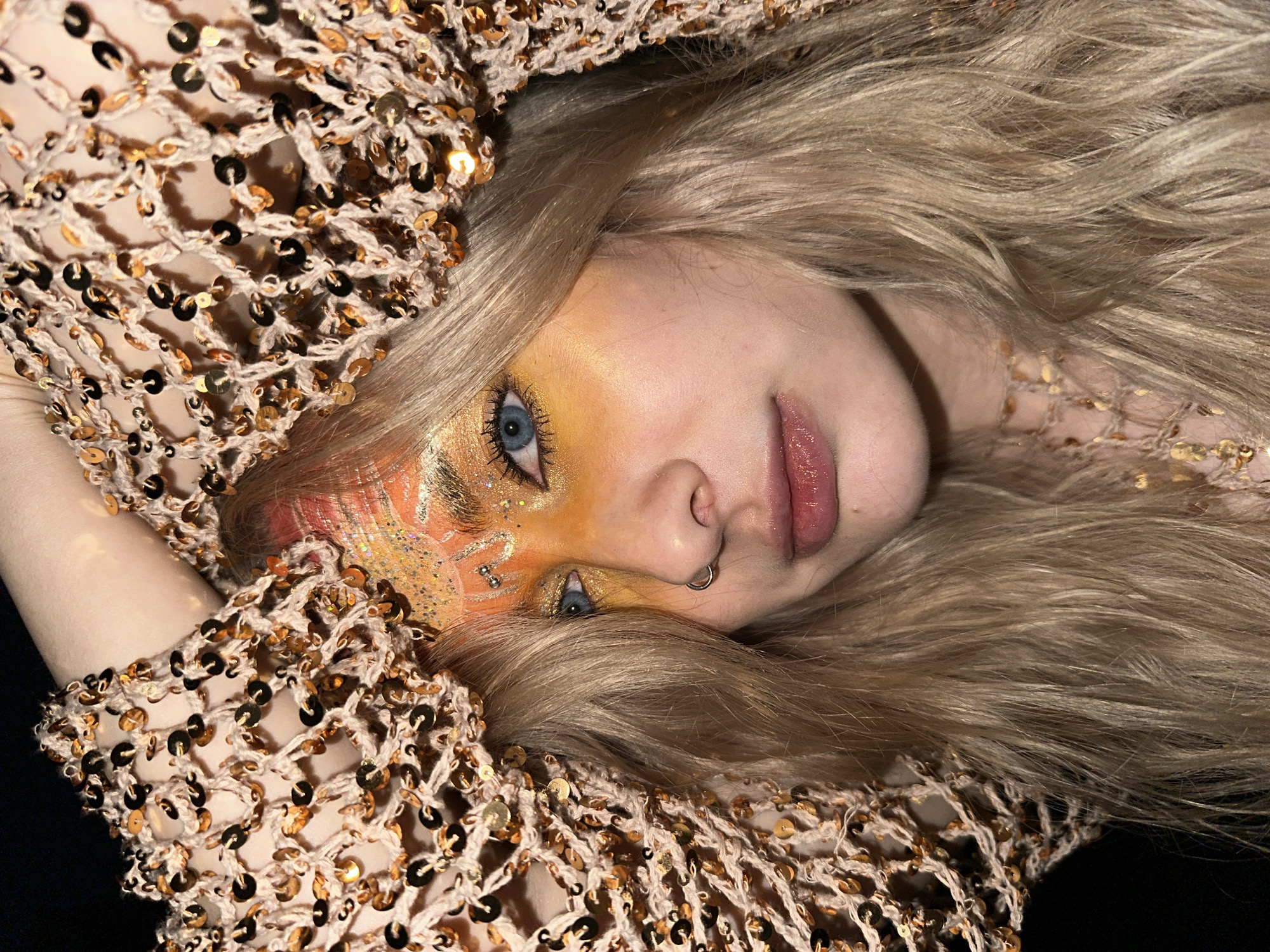 A close-up, angled view depicts a person with light blonde hair and blue eyes, lying down. The person has theatrical makeup featuring shades of orange and glitter around one eye, and a nose ring. The person is covered in a beige, sequined, loosely-woven fabric. The background appears to be dark, possibly black. The location is not discernible as Chaikovsky, Russia.