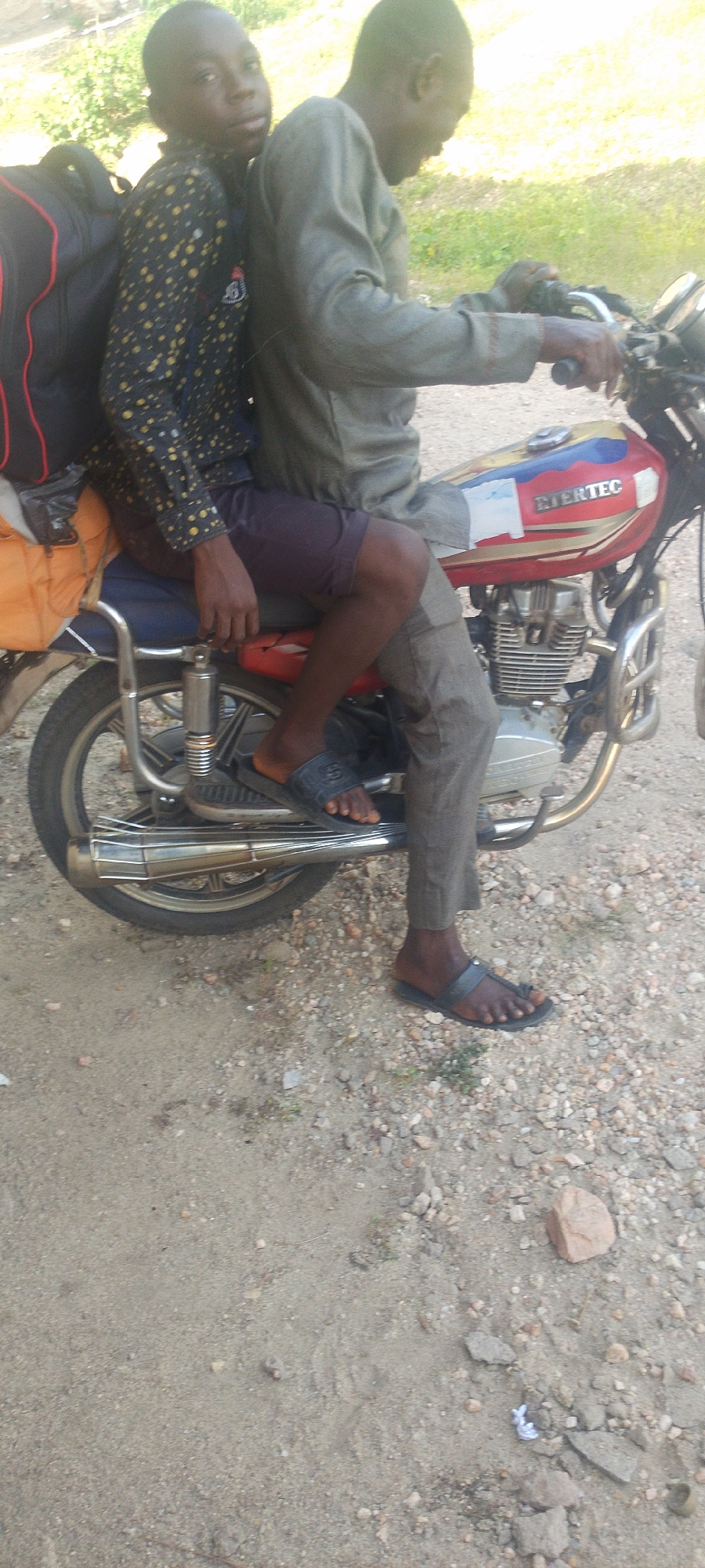 Two African individuals are seated on a red and white motorcycle labeled "BIERTEC." The rider, wearing a long-sleeved grey shirt and grey pants, operates the motorcycle. The passenger, wearing a black and yellow patterned long-sleeved shirt and short pants, sits behind the rider with a large backpack. Both are wearing sandals. The motorcycle is parked on a dirt road. The location is consistent with a rural or semi-rural area in Lokoja, Nigeria. Vegetation is visible in the background.