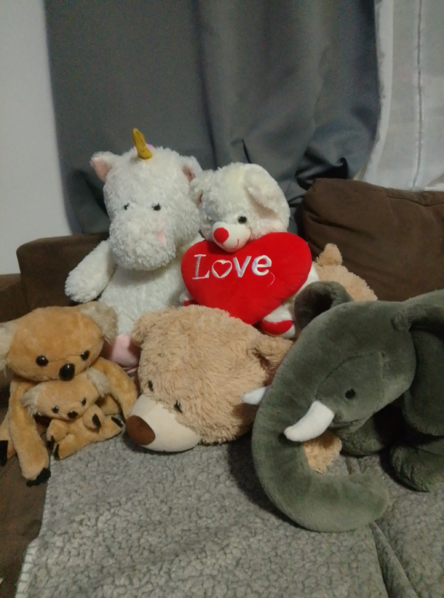 A group of stuffed animals is arranged on a couch. The stuffed animals include a white unicorn with a yellow horn, a white dog with a red heart that says "Love", a brown koala with a smaller koala on its lap, a brown bear, and a green elephant. The couch is covered with a grey, fuzzy blanket. The background consists of a grey curtain. The image was taken in Wedel, Germany.