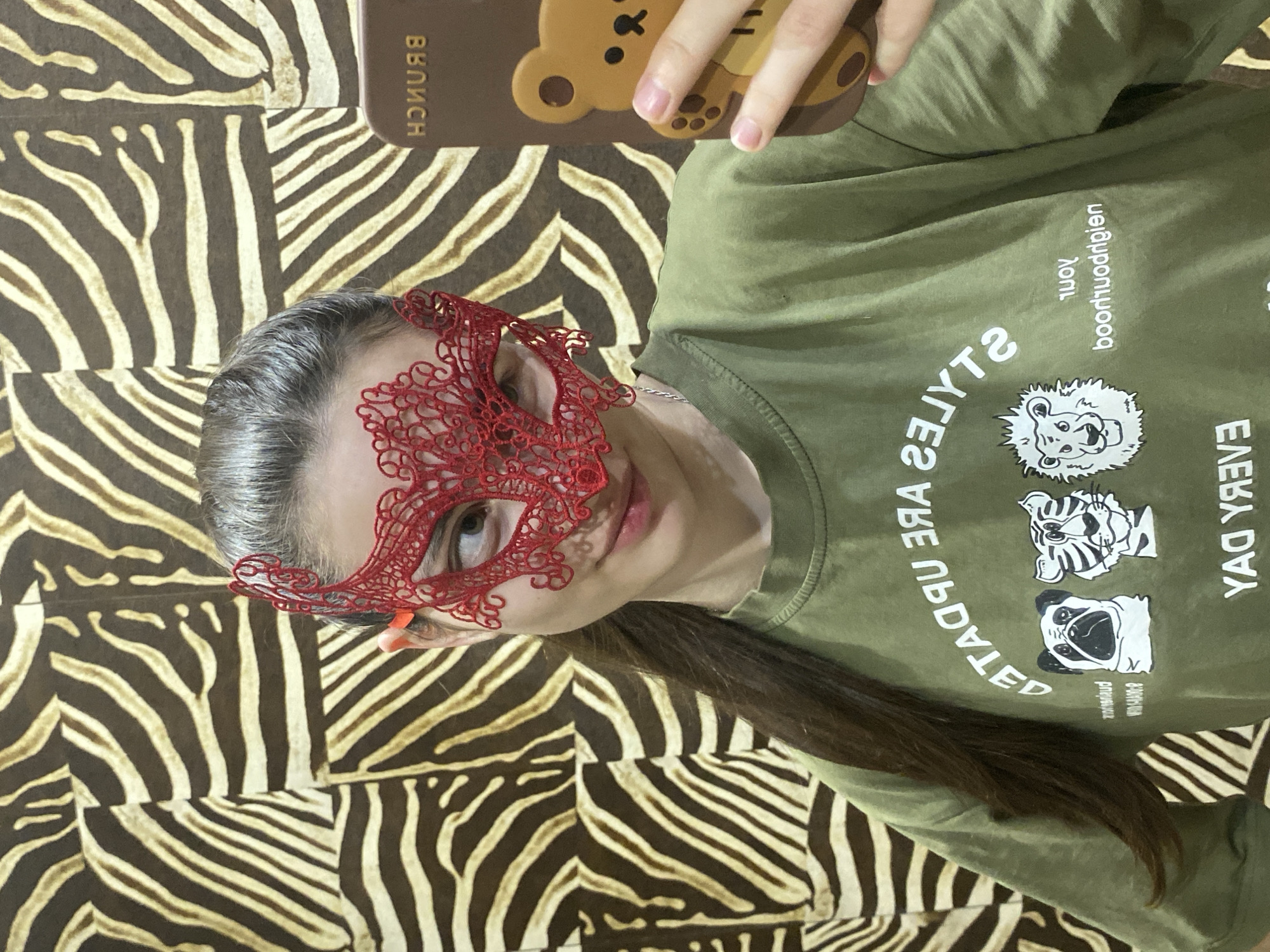 A person is captured in a selfie, holding a phone to take the picture, in what appears to be an indoor setting in Votkinsk, Russia. The image is rotated clockwise, showing the person's upper body and head from a slightly elevated angle. She is wearing a red lace masquerade mask over her eyes, with a hint of a smile on her visible lower face. Her long, dark hair cascades down her back and shoulder.
She wears an olive green t-shirt adorned with white graphics of animal faces: a lion, a tiger, and a pug. Partially visible text on the shirt, reversed due to reflection, includes phrases like "EVERY DAY," "goodneighborhood," and "STYLES ARE UPDATED." She holds a brown phone case with a cartoon bear design, and the word "BRUNCH" is visible on the case, also reversed. The background features a striking zebra-print pattern, possibly on a wall or piece of furniture. The overall mood is casual and playful.