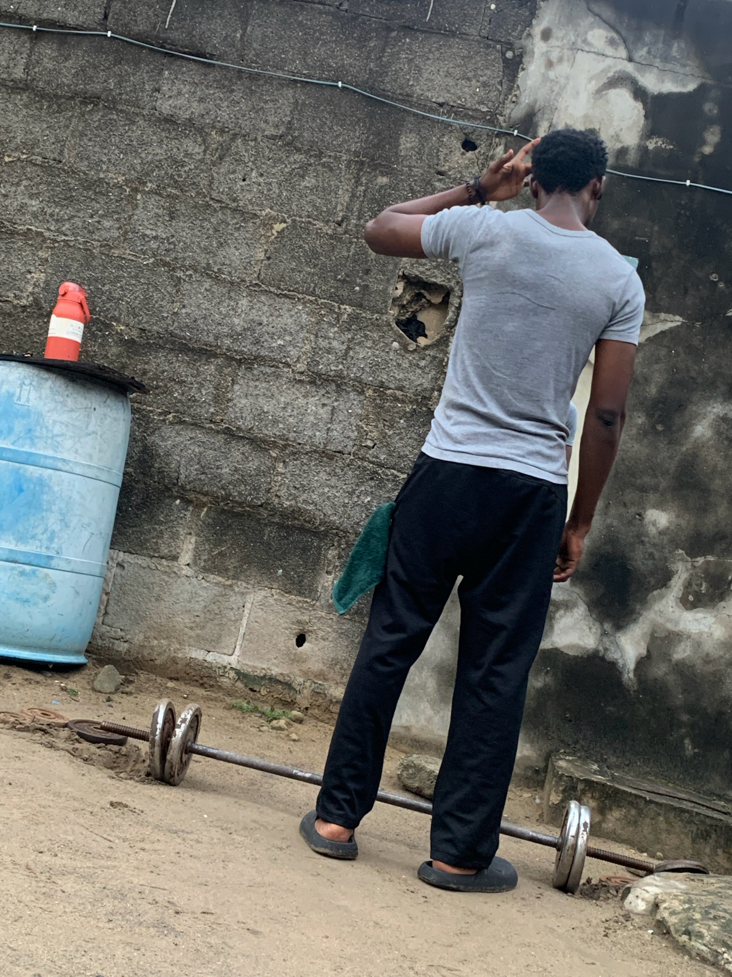 A young Black male stands with his back to the camera, facing a weathered concrete wall in Ado, Nigeria. He is wearing a grey t-shirt and black pants, with a dark green cloth tucked into his waistband. His right hand is raised to his forehead in a gesture that could indicate contemplation or shielding his eyes. In the foreground, a barbell with two weight plates on each side lies on the dusty ground. To the left, a large blue plastic barrel with a red and white traffic cone on top is positioned against the wall. The environment appears to be an outdoor urban space, likely a backyard or alley, characterized by its rough textures and utilitarian objects.