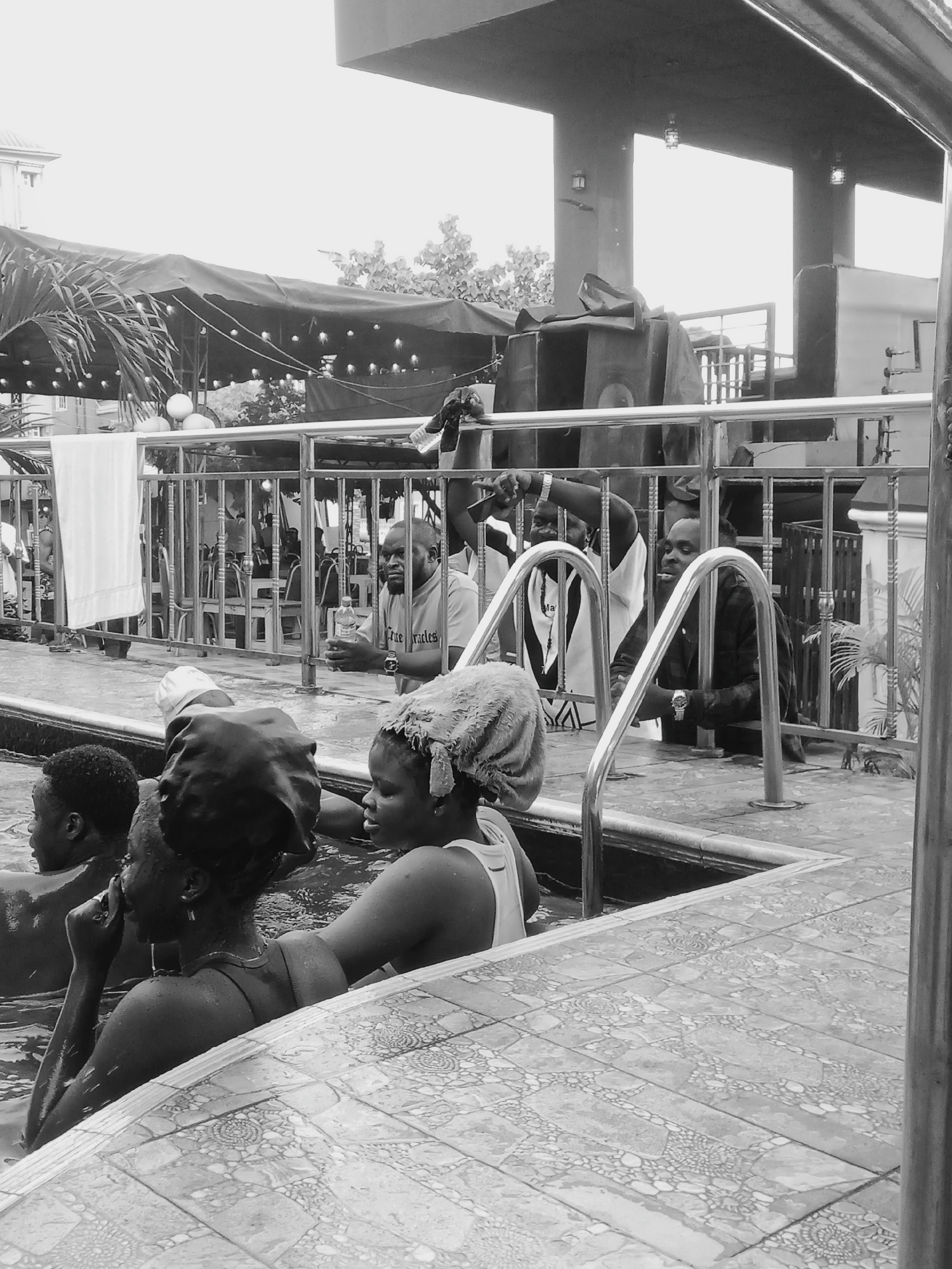 The image depicts a group of people in and around a swimming pool in Onitsha, Nigeria.
Several individuals are in the water, up to their shoulders. One person has a towel wrapped around their head. Another appears to be speaking on a phone.
A metal railing separates the pool area from a slightly elevated patio. On the patio, several men are standing or leaning against the railing. Some are holding drinks.
Behind the patio, there is a covered area with strung lights. A multi-story building is visible in the background. The ground surrounding the pool is tiled.