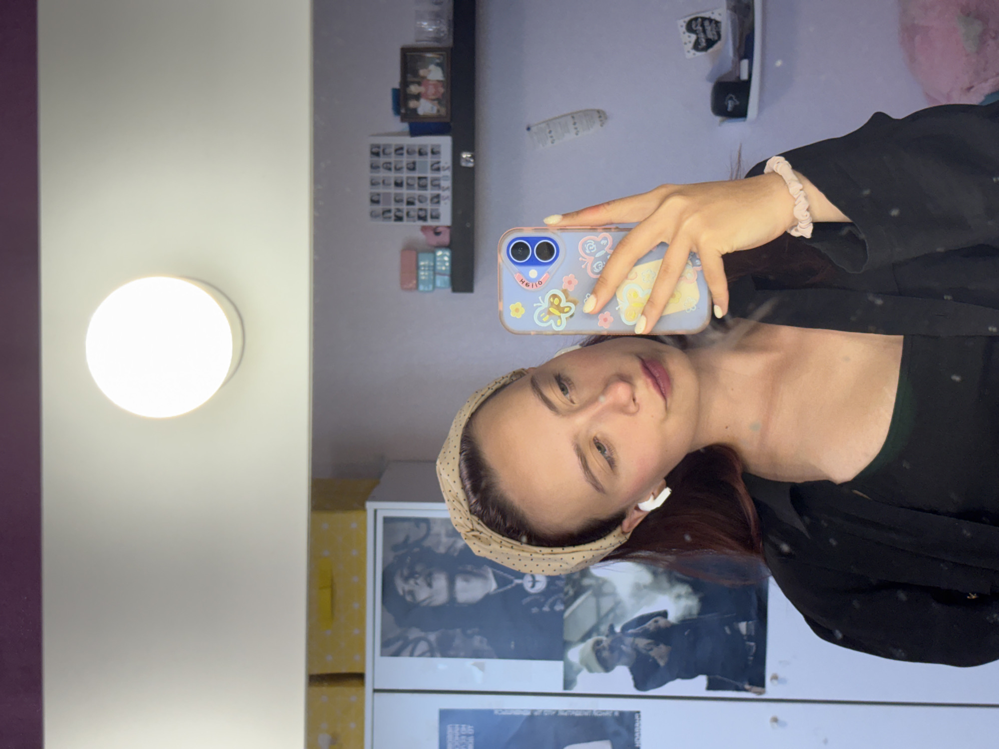 In a dimly lit room, a young woman with red hair, wearing a black shirt and a beige headband, takes a selfie in a mirror. She has white earbuds in her ears and is holding a light blue phone with a butterfly design. Behind her, a shelf holds various items, including a framed family photo and a collection of smaller framed pictures. To her left, posters of musicians are visible on a cabinet. The overall setting appears to be a personal space, possibly a bedroom or dressing area. The image does not provide specific information about the location within the city of Прикамский, Russia.