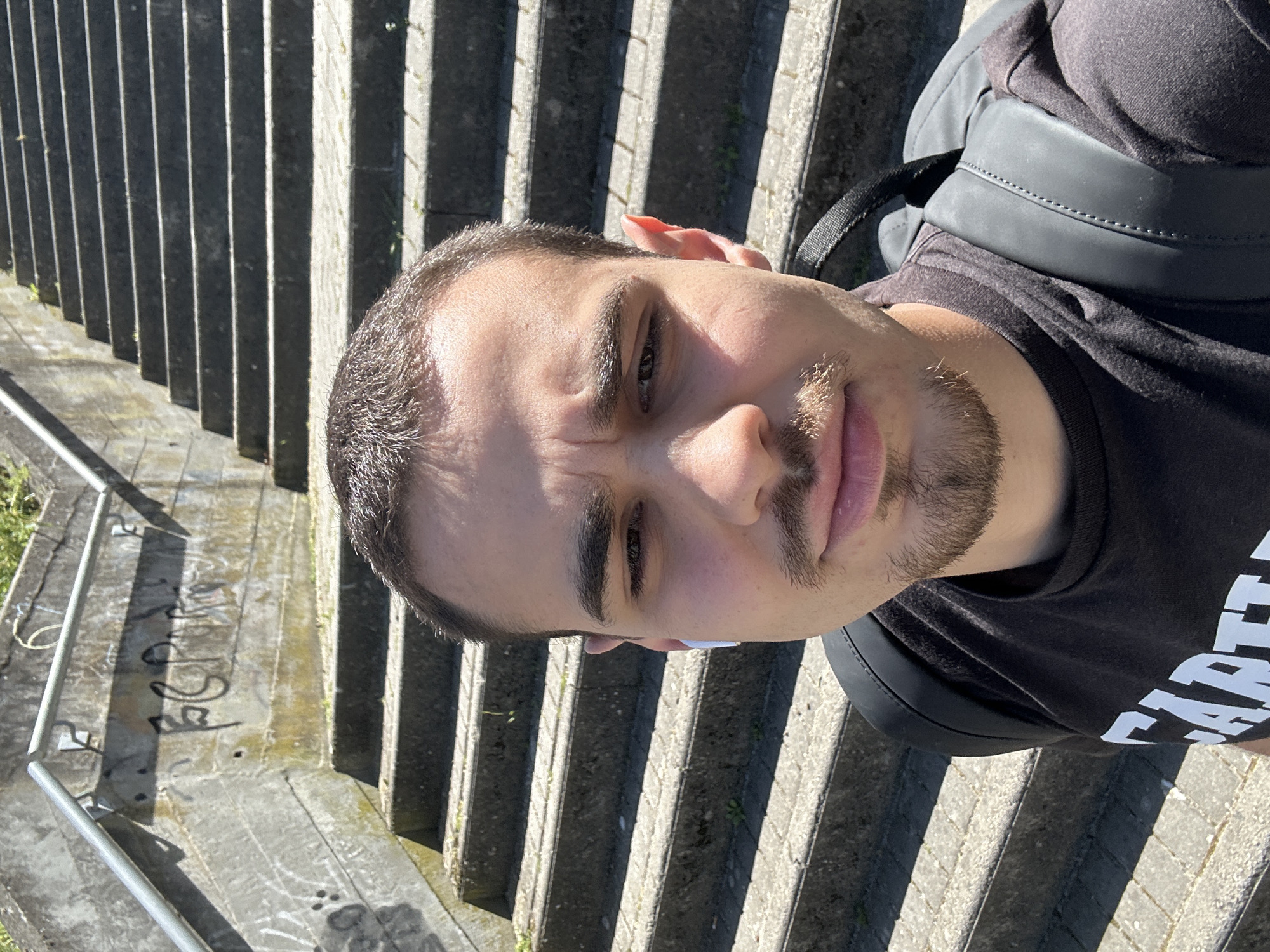 The image shows a man standing on a set of concrete stairs. He is wearing a black T-shirt and a black backpack. The stairs appear to be outdoors, possibly in a public area. There is graffiti on the adjacent ground. The location is the Mirandela stairs in Portugal.