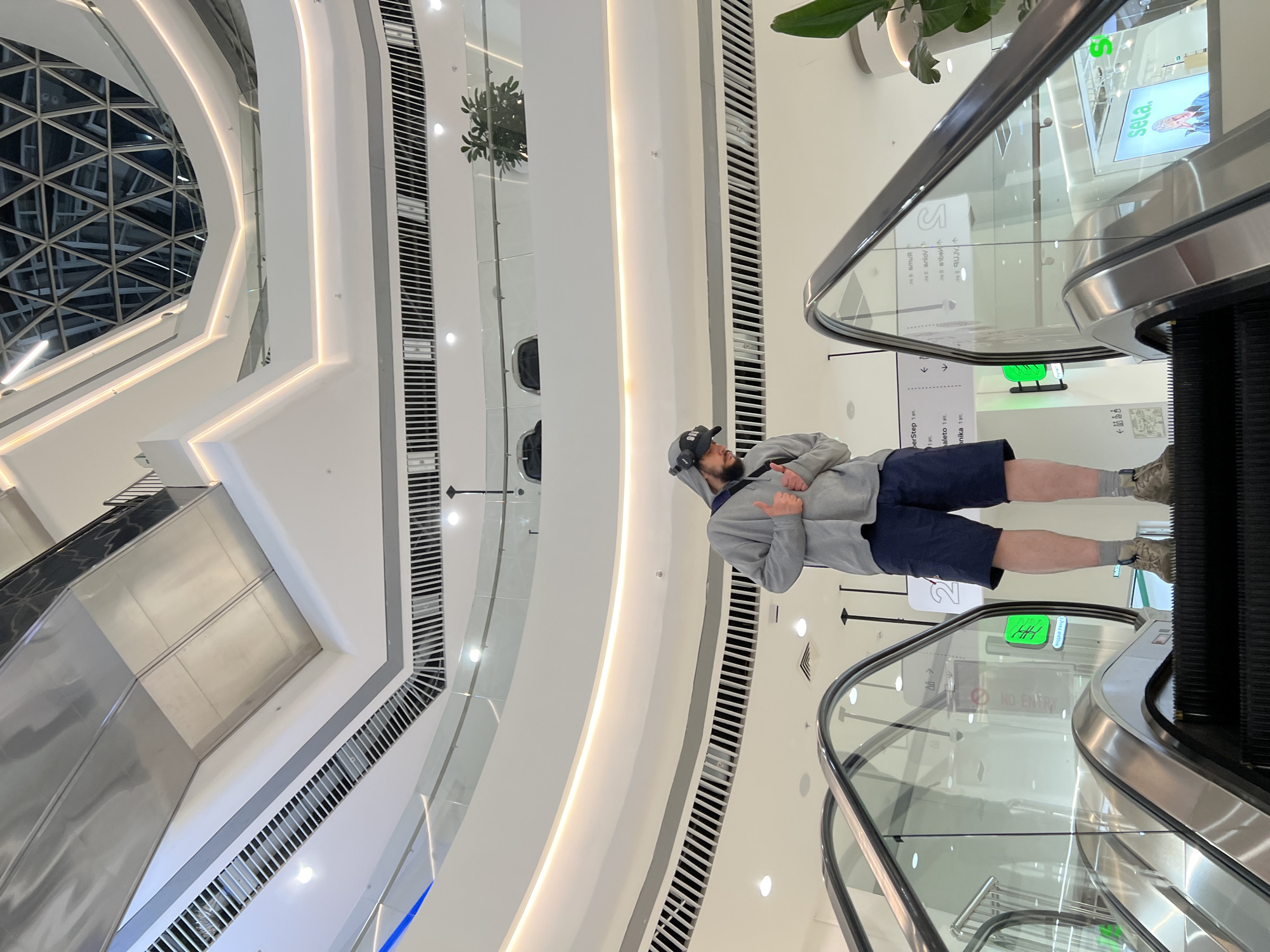 The image displays the interior of a modern, multi-story shopping mall or commercial complex located in Khabarovsk, Russia. The setting is characterized by clean, brightly lit white walls with elegant curved lines and integrated strip lighting. A prominent feature in the upper left is a large, geometric glass dome or skylight structure, suggesting a contemporary architectural style. Multiple escalators with clear glass railings and polished metal handrails connect the different levels of the building. Decorative green plants are visible in planters on the upper floors.
The primary subject is a man standing on an escalator. He is dressed casually in a grey hooded sweatshirt, dark blue shorts, and camouflage-patterned sneakers with grey socks. A black baseball cap is on his head, and black headphones rest around his neck. He has a beard and is looking upwards and slightly to his right. With both hands, he is making a "shaka" or "hang loose" gesture, suggesting a relaxed, positive, or playful mood, possibly while posing for the camera.
Several signs and text elements are visible throughout the mall. A "NO ENTRY" sign in a red circle is seen near an escalator. Other text includes what appear to be store names or floor indicators in Cyrillic, such as "berStep 1эт." (likely "SuperStep 1st floor"), "aleto 1эт." ("Paleto 1st floor"), and "onika 1эт." ("Monika 1st floor"). A large advertisement features the brand "sela.". Other discernible Cyrillic words include "теватия", "anlov", "sabel", "Видия ту", and "Выхо" (likely part of "Выход", meaning "Exit") accompanied by a left arrow. The scene is brightly lit by artificial light, indicating an indoor environment where the time of day and external weather are not relevant.